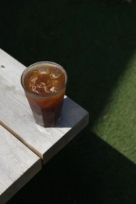 A glass of iced coffee on a sunlit wooden surface with grass in the background.