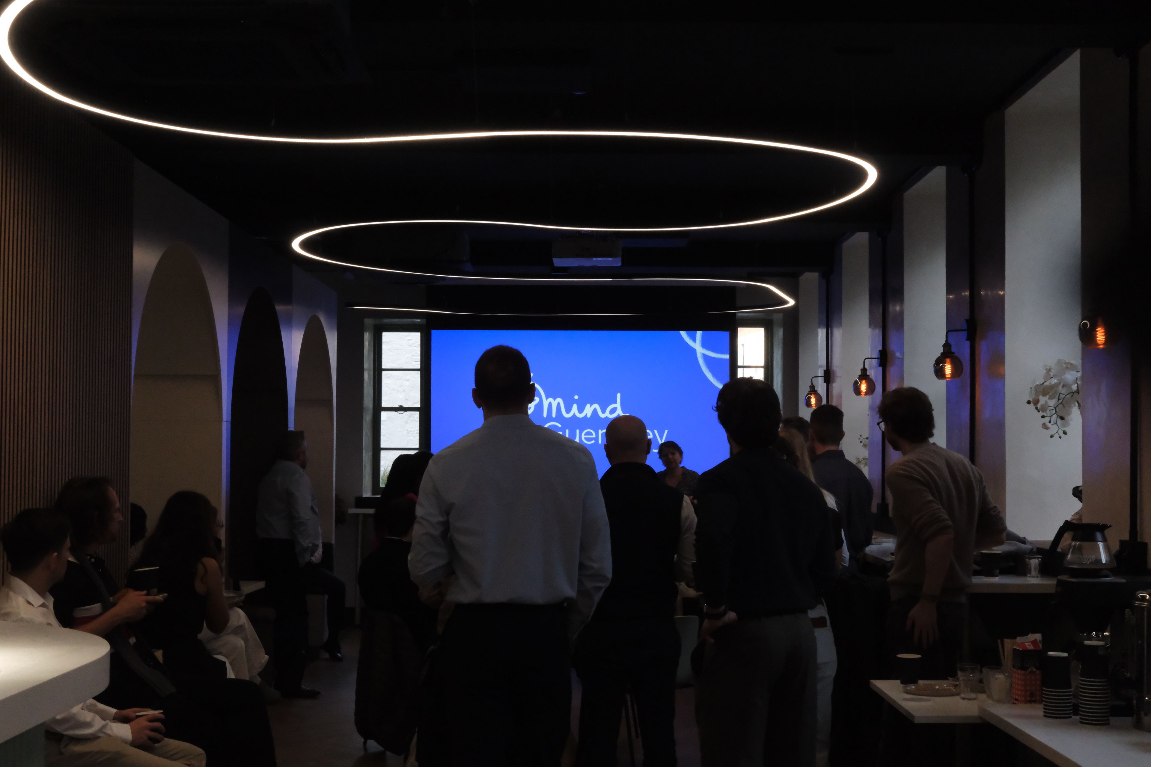 People gathered in a modern room with curved ceiling lights and a large screen displaying "Mind Guernsey."