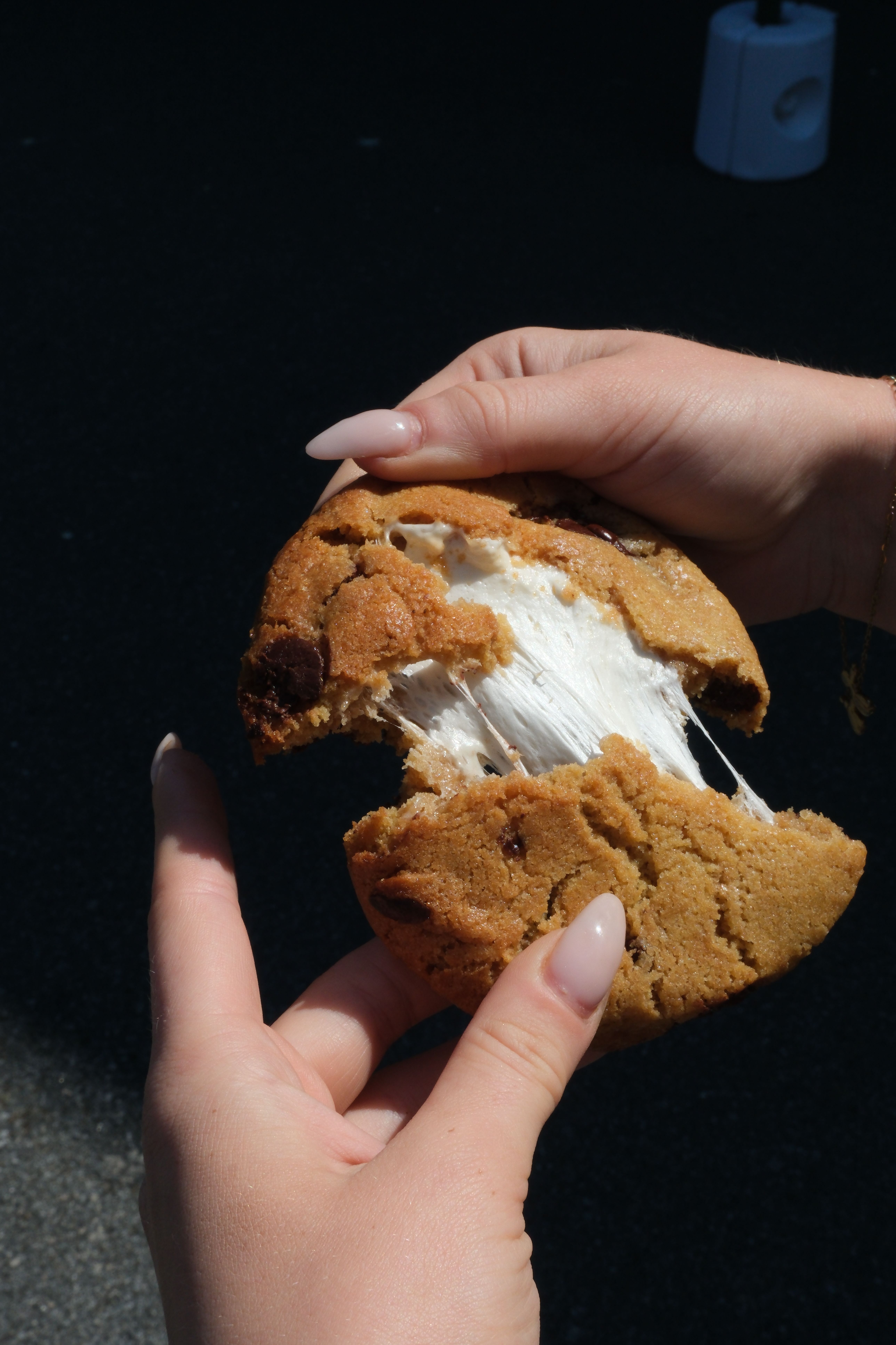 Hands breaking a chocolate chip cookie in half, revealing gooey, stretching marshmallow.