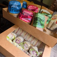 Two catering boxes: the top with bags of Brown Bag Crisps and wraps, the bottom with assorted sandwiches and wraps.