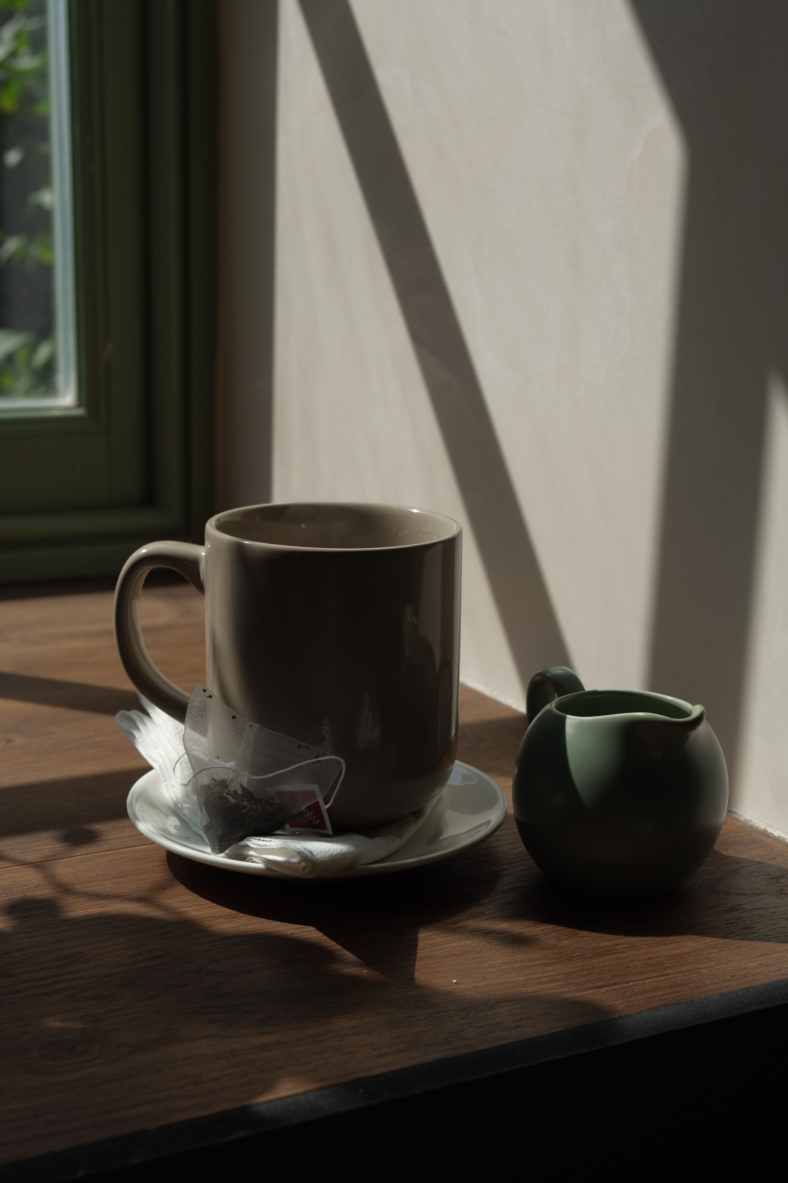 A beige mug with a tea bag on a saucer, and a green pitcher, sit on a wooden surface with strong sunlight and diagonal shadows.