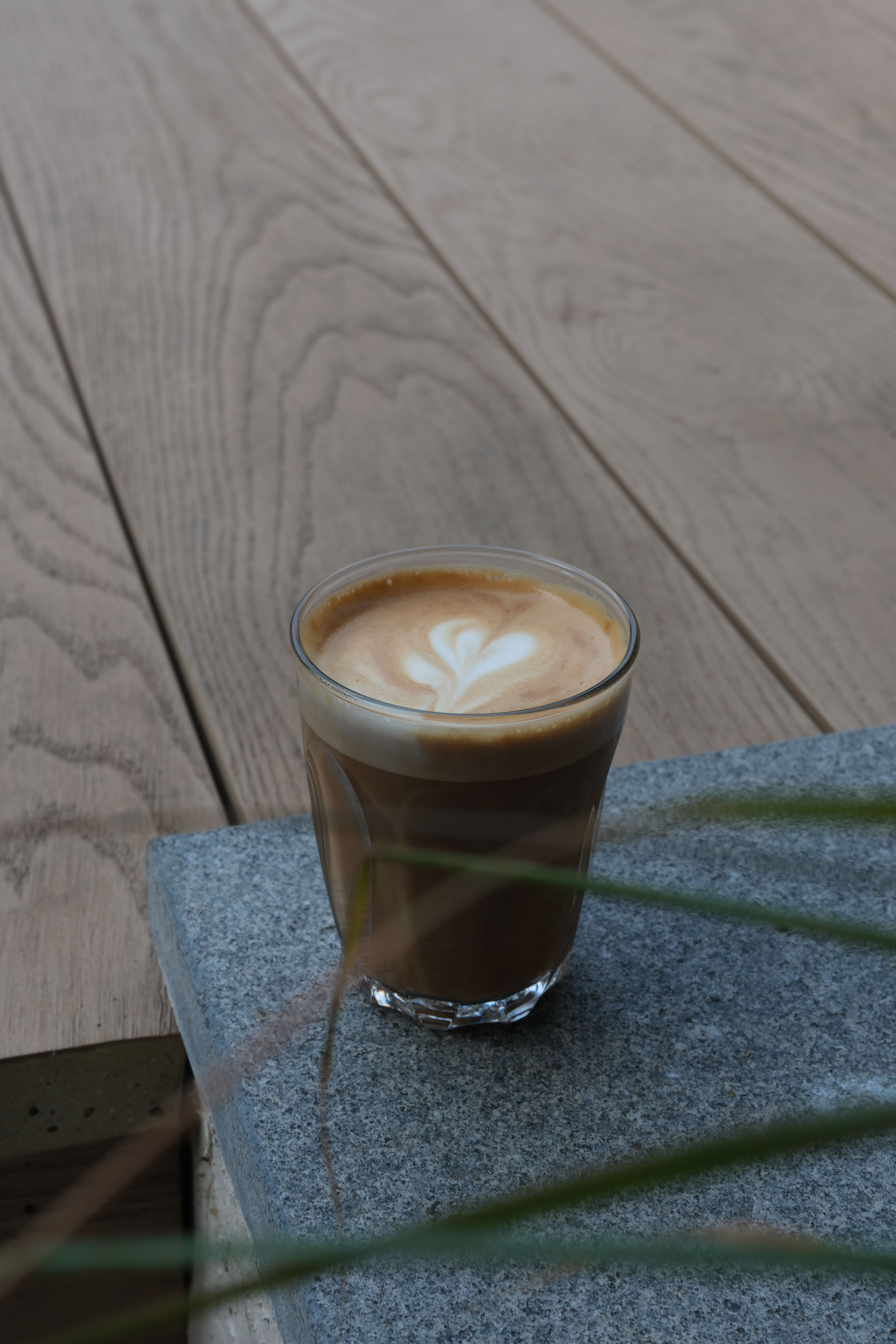 A latte with heart art in a glass on grey stone and a wooden deck.