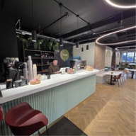 A barista stands behind a modern cafe counter with a light green fluted front and "Camila's" sign, in a space with herringbone flooring and varied seating.