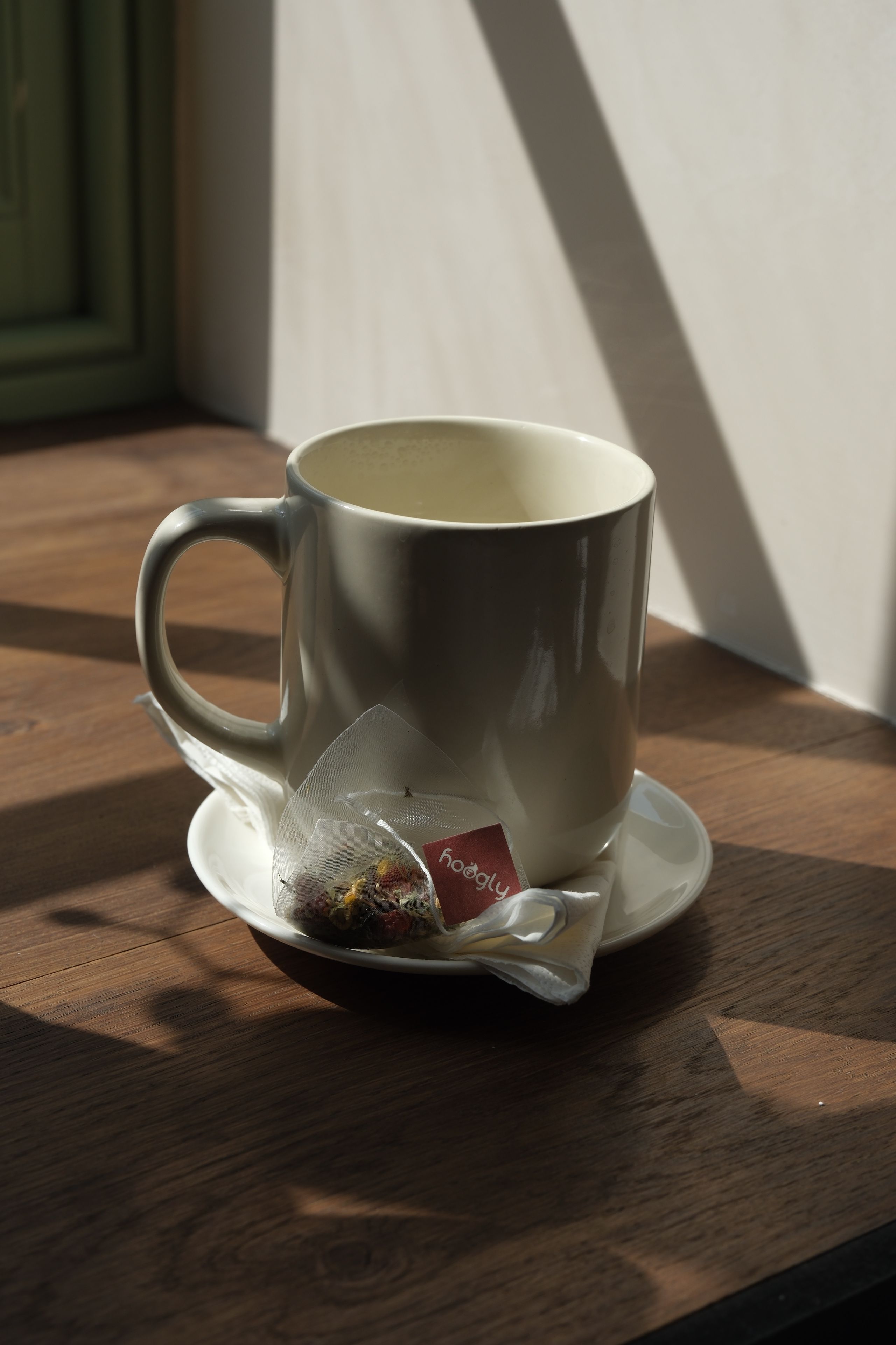 A white mug and a Hoogly brand tea bag on a saucer, resting on a wooden table in sunlight.