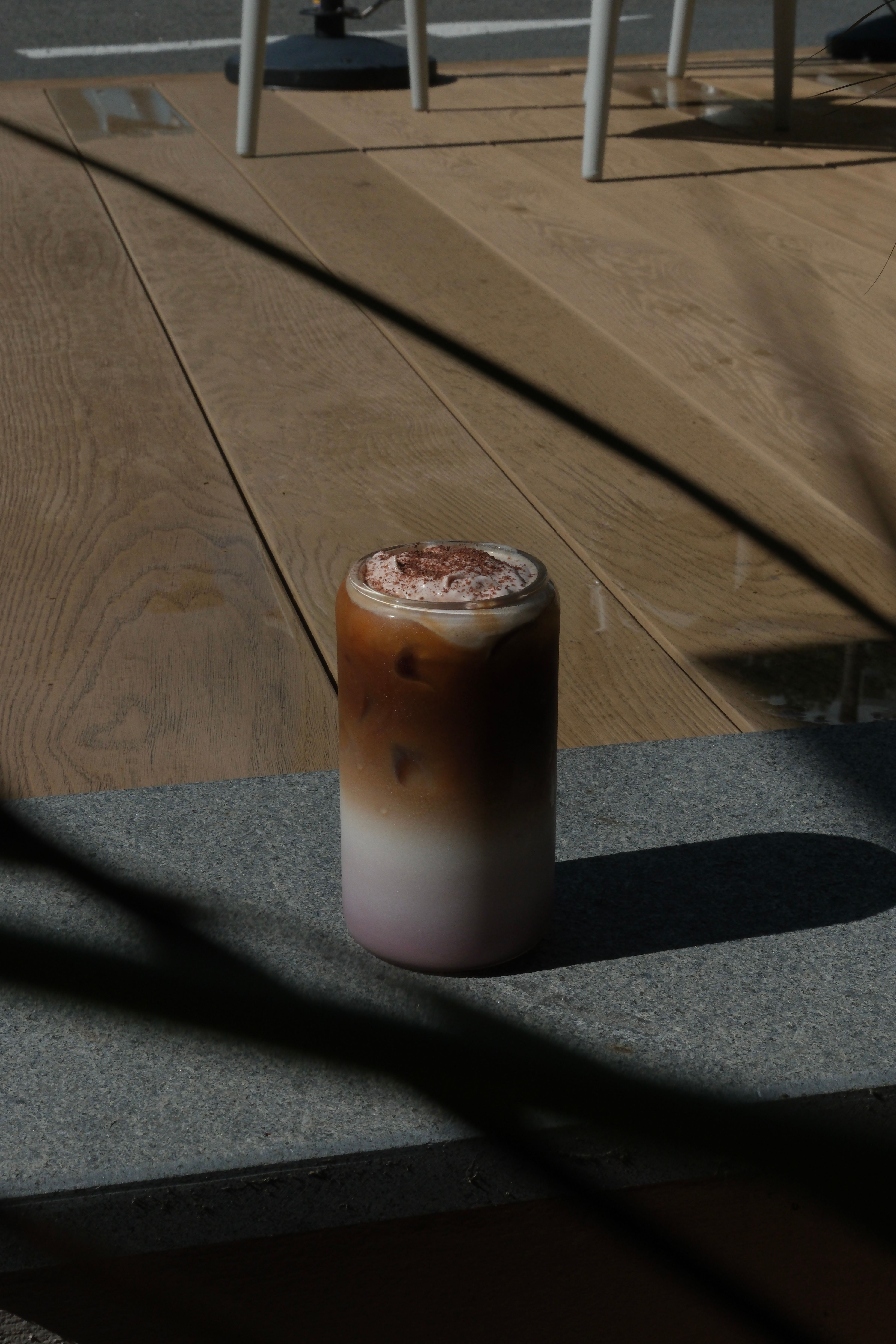 A layered iced drink with pink, white, and brown layers, topped with whipped cream and cocoa powder, on a gray surface in sunlight.
