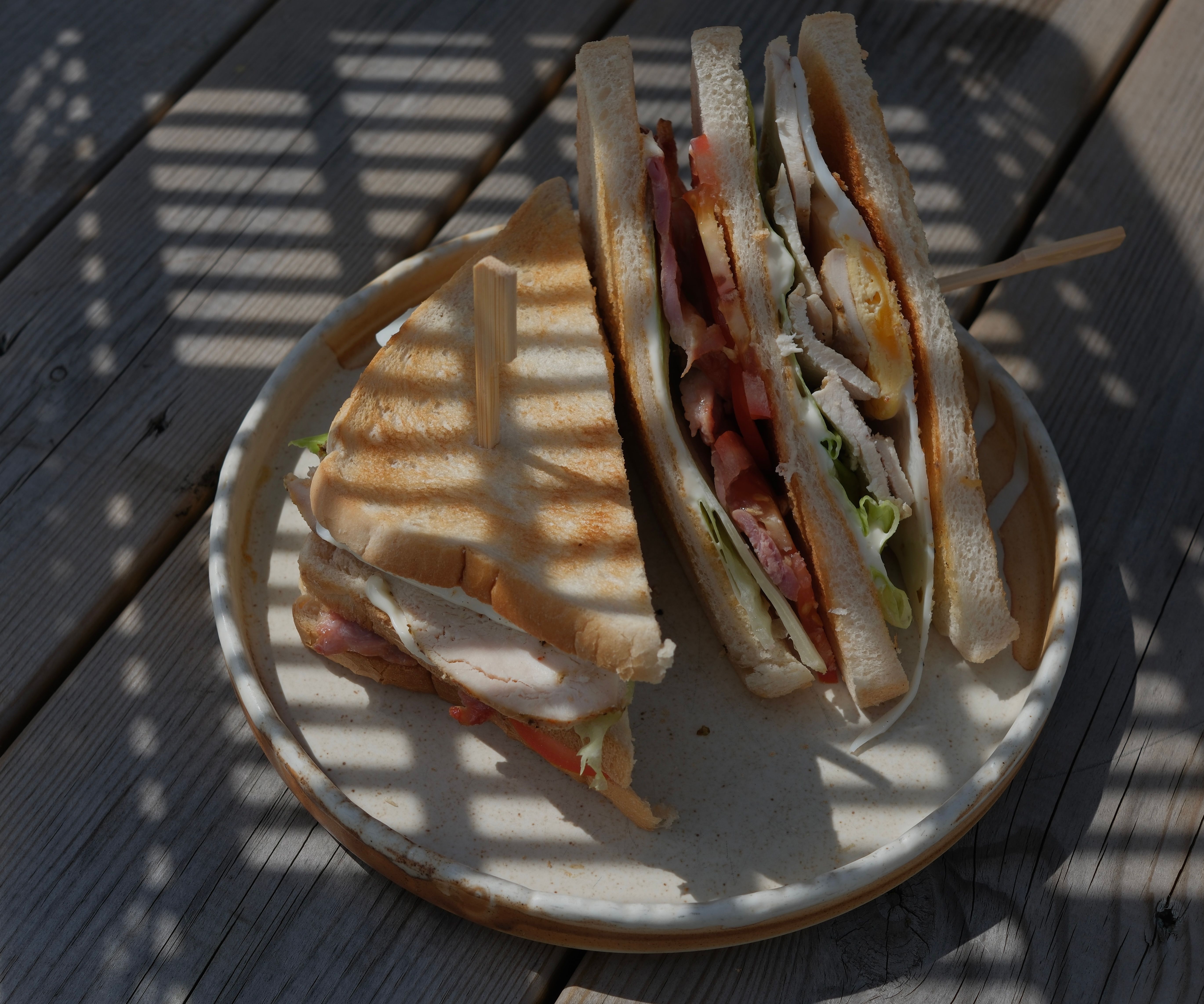 A club sandwich with bacon, chicken, egg, lettuce, and tomato, on a plate on a shadowed wooden table.