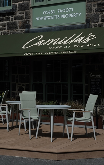 Camilla's Cafe at the Mill exterior with a green awning, outdoor tables, light green chairs, and a "WATTS.PROPERTY" sign on the stone building.