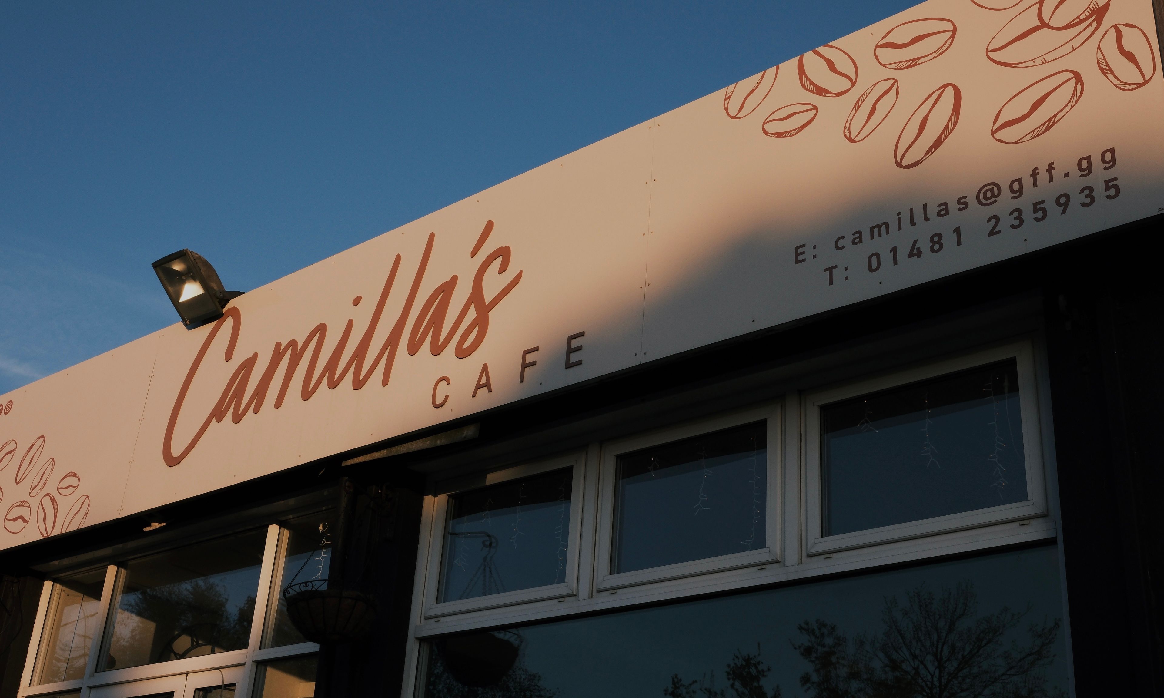 Camilla's Cafe sign on a building facade, featuring coffee bean illustrations and contact information.