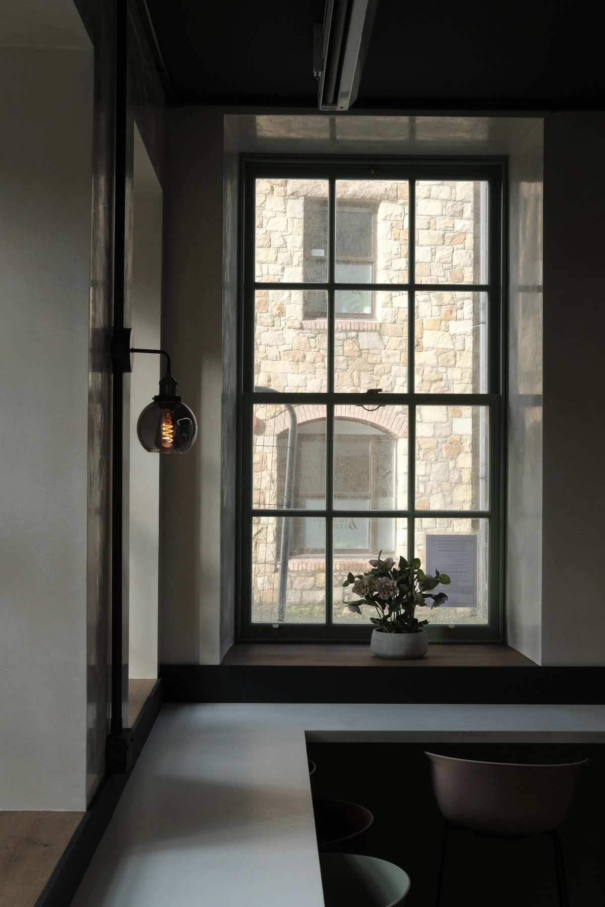 A green-framed window looks out to a stone building, with a potted plant on the sill and a wall lamp to the left.