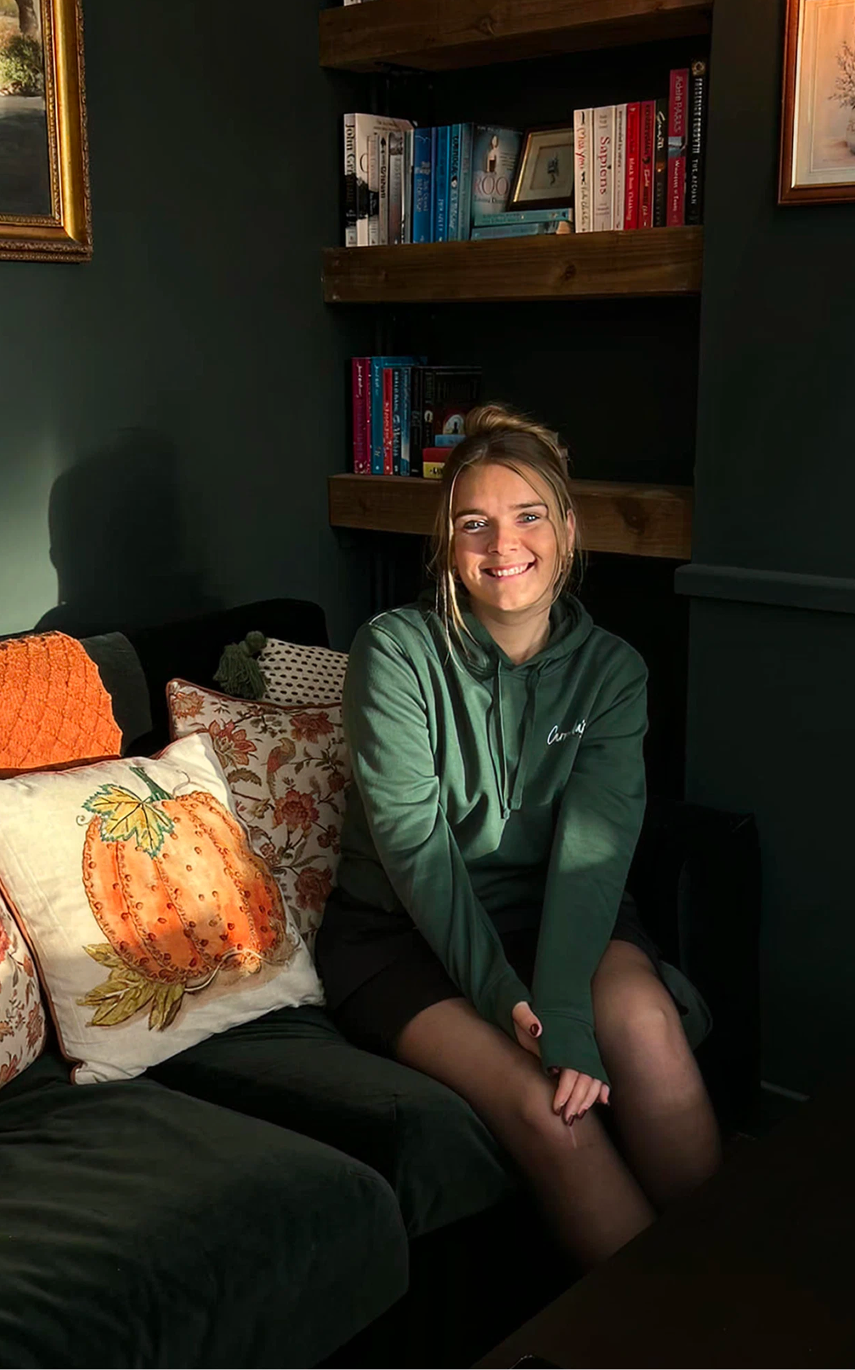 A smiling woman in a green hoodie sits on a couch with a pumpkin pillow, bathed in sunlight, with bookshelves behind her.