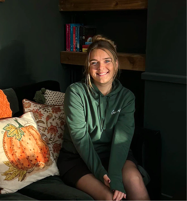 Smiling woman in a green hoodie sitting on a couch with a pumpkin pillow, lit by sunlight.