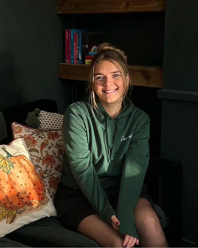 Smiling woman in a green hoodie sitting on a couch with a pumpkin pillow, lit by sunlight.