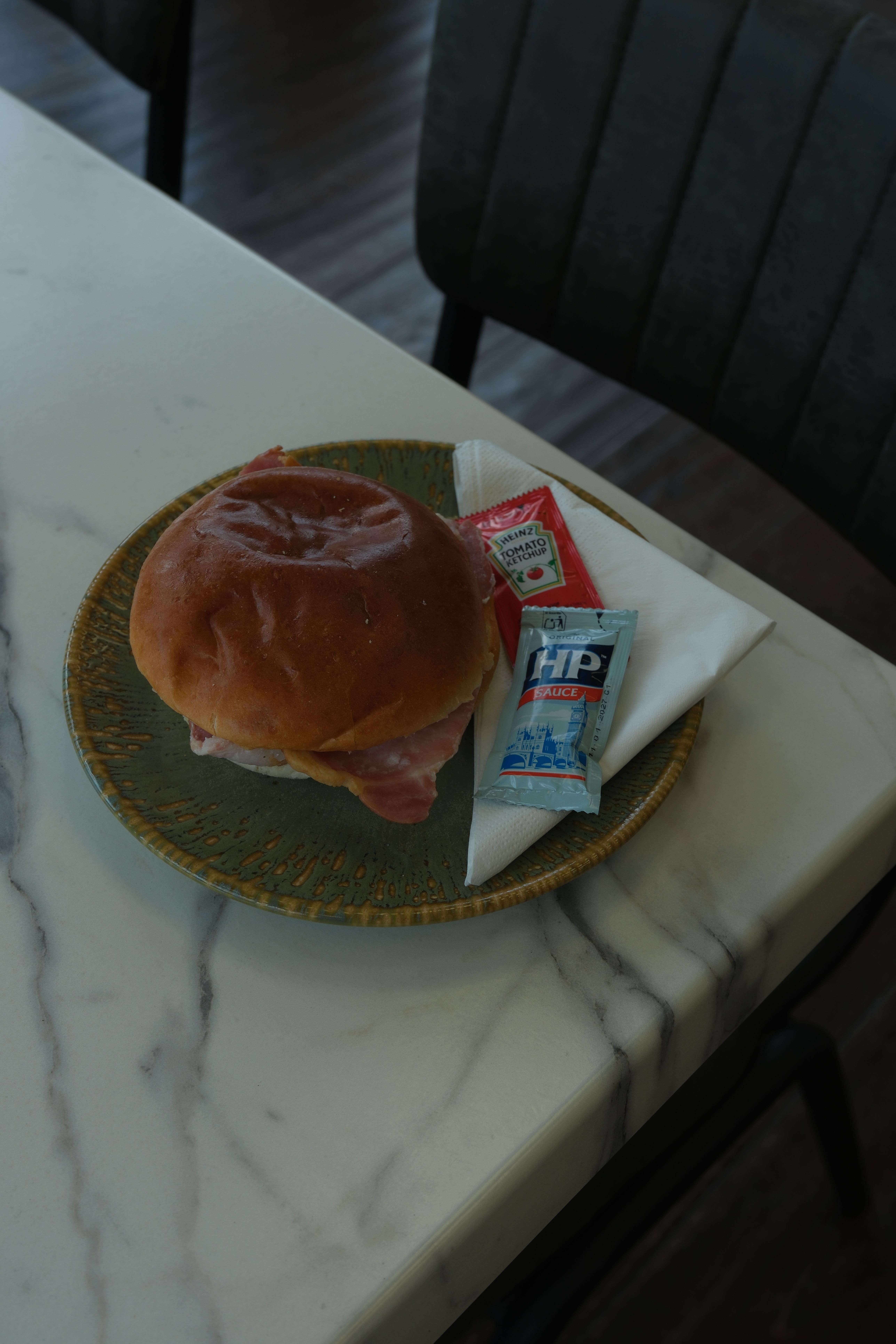 A bacon roll on a green plate with ketchup and HP sauce packets on a marble table.