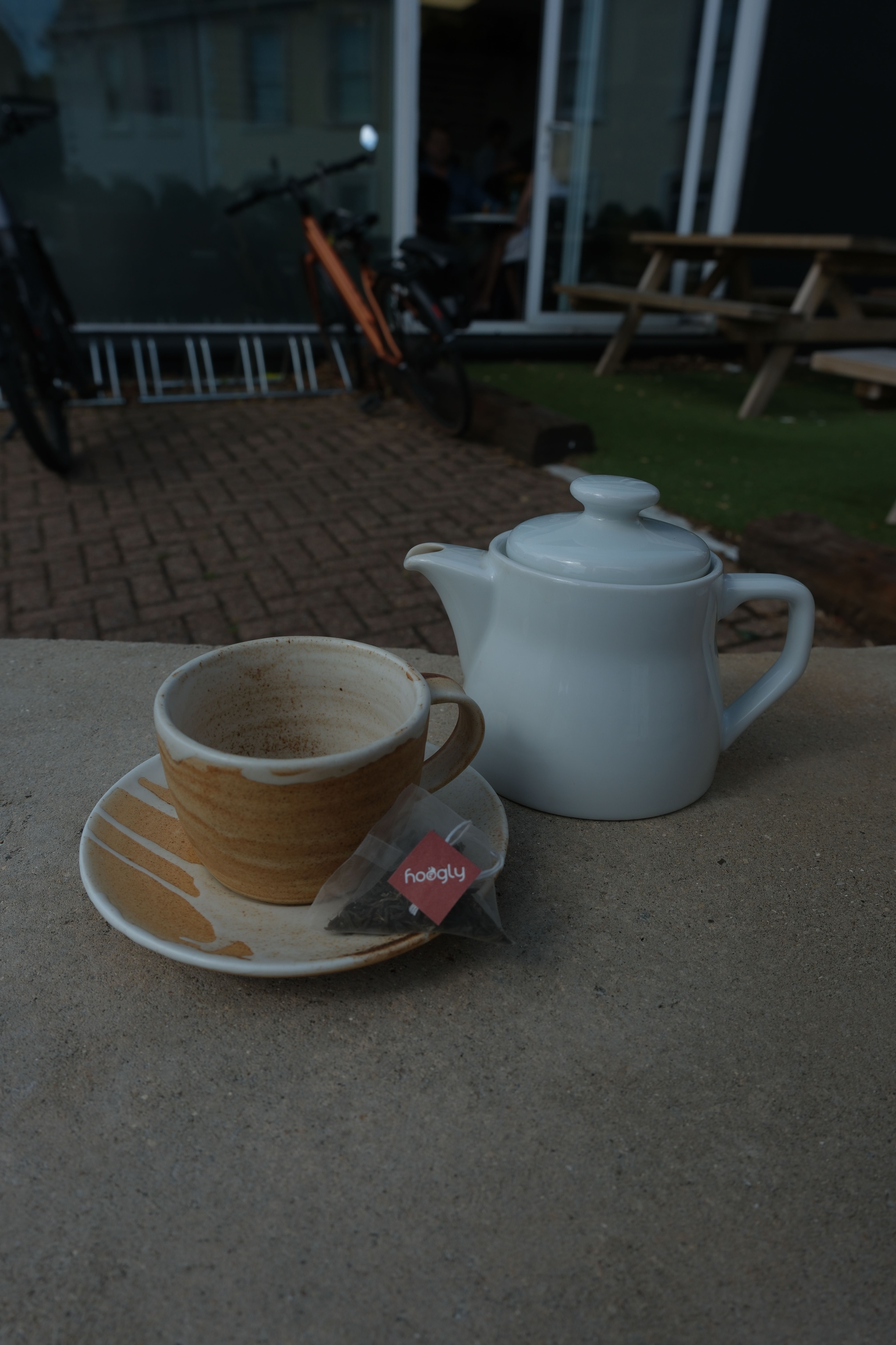 Rustic ceramic teacup and saucer, a white teapot, and a Hoogly tea bag on a concrete surface.