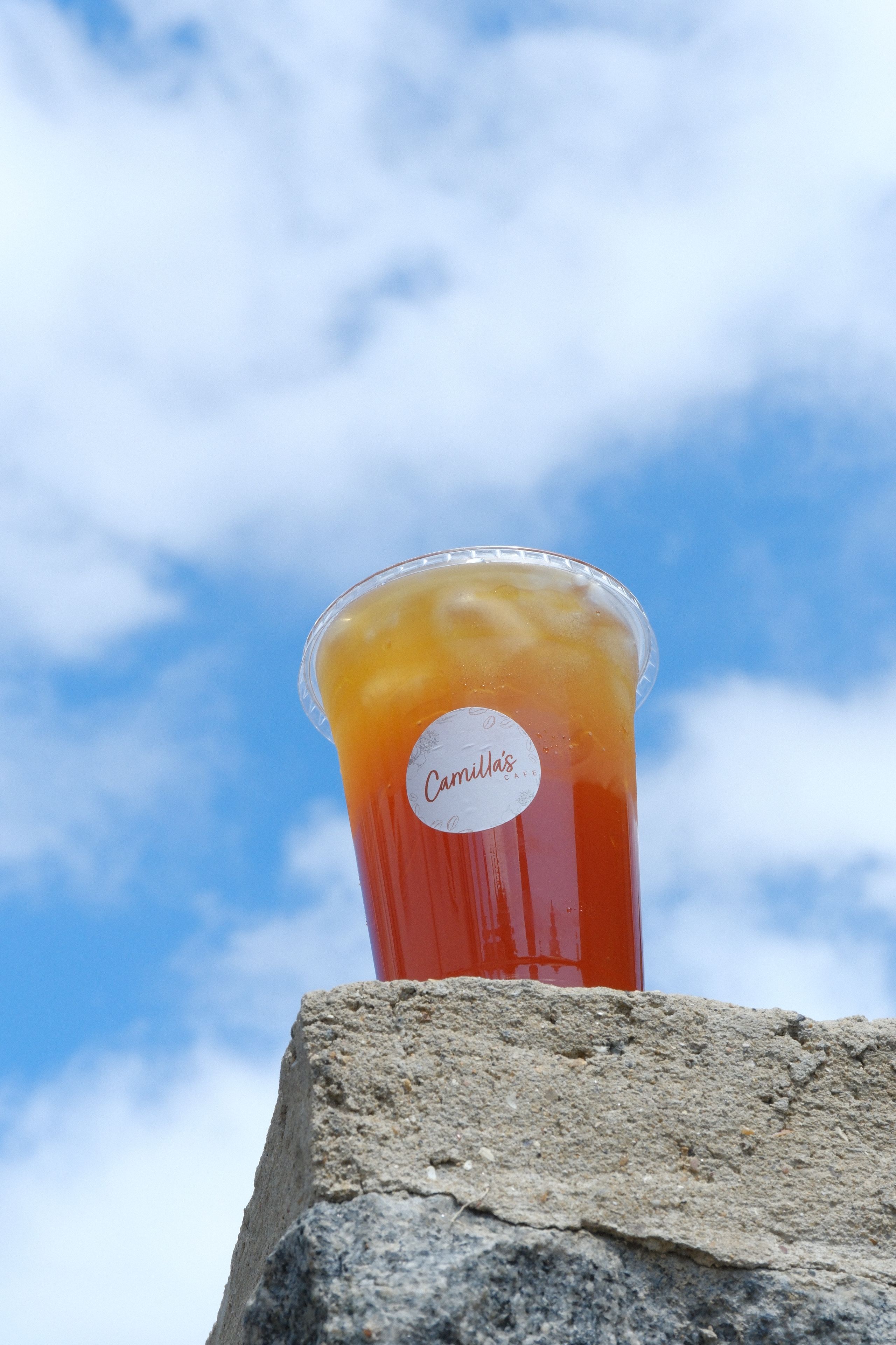 A clear plastic cup with a layered orange and red iced drink, labeled "Camilla's", sits on a stone wall under a blue sky with clouds.