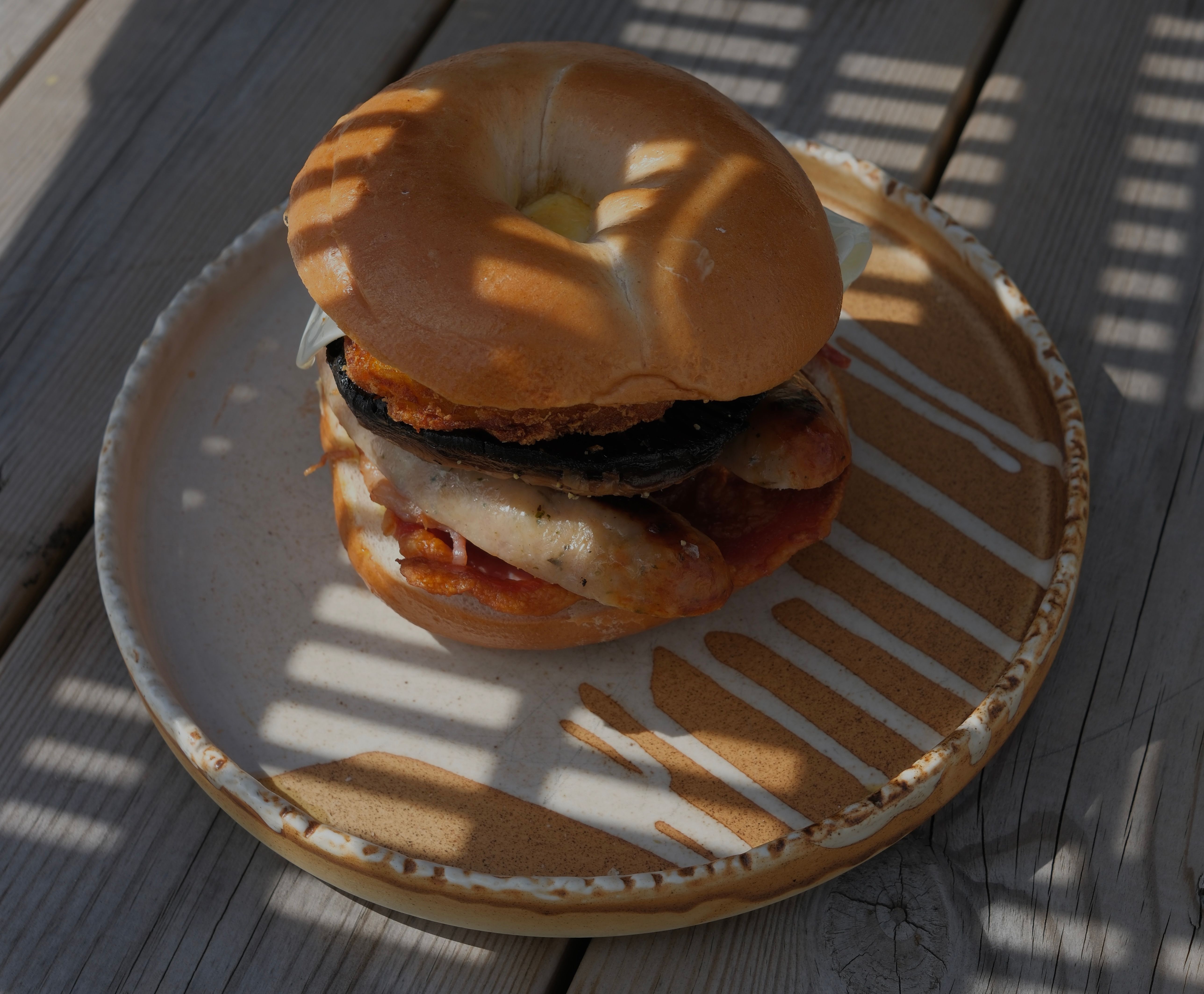A bagel sandwich filled with sausage, mushroom, and bacon, resting on a decorative plate on a wooden table.