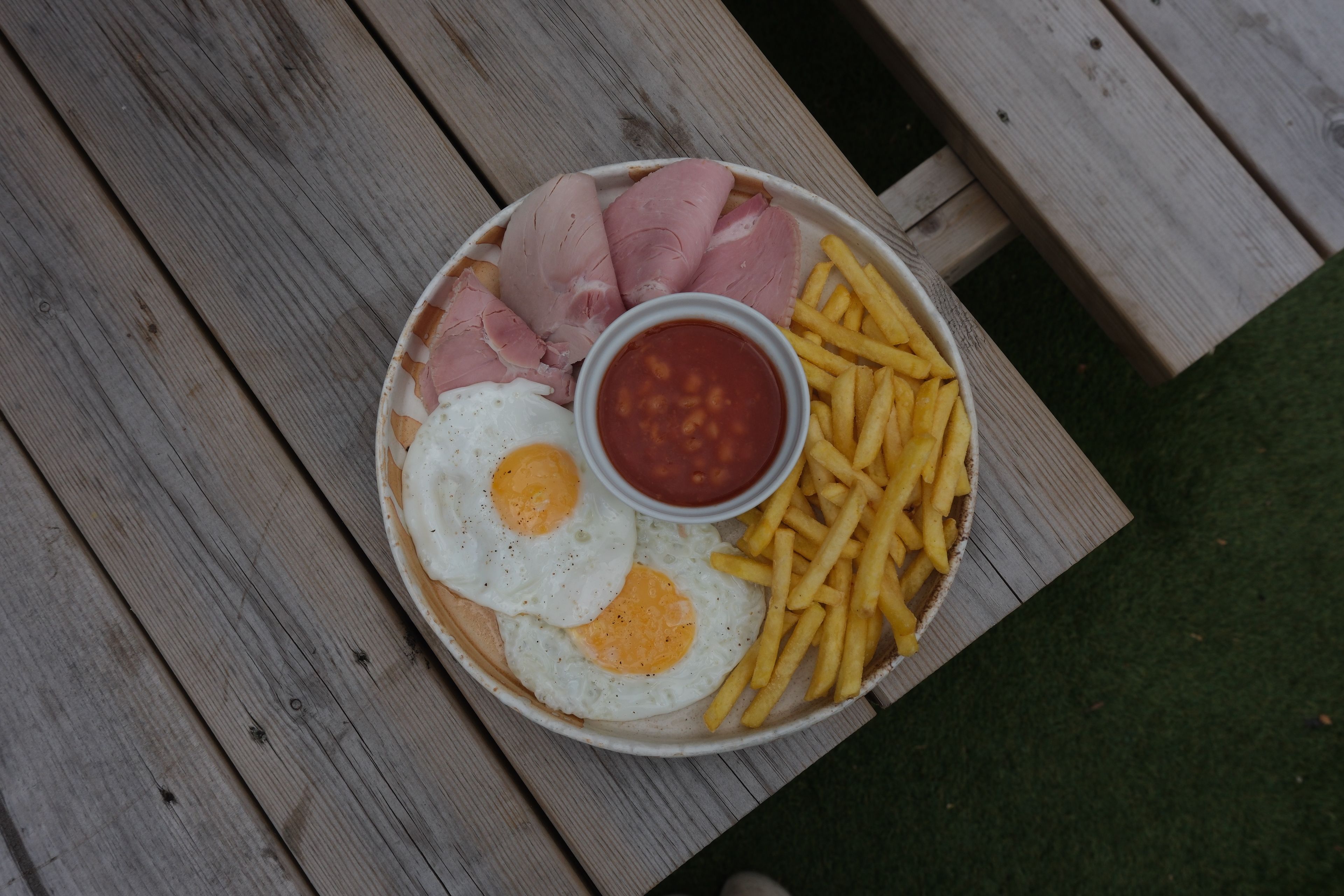 A plate of fried eggs, ham, baked beans, and french fries on a wooden table.