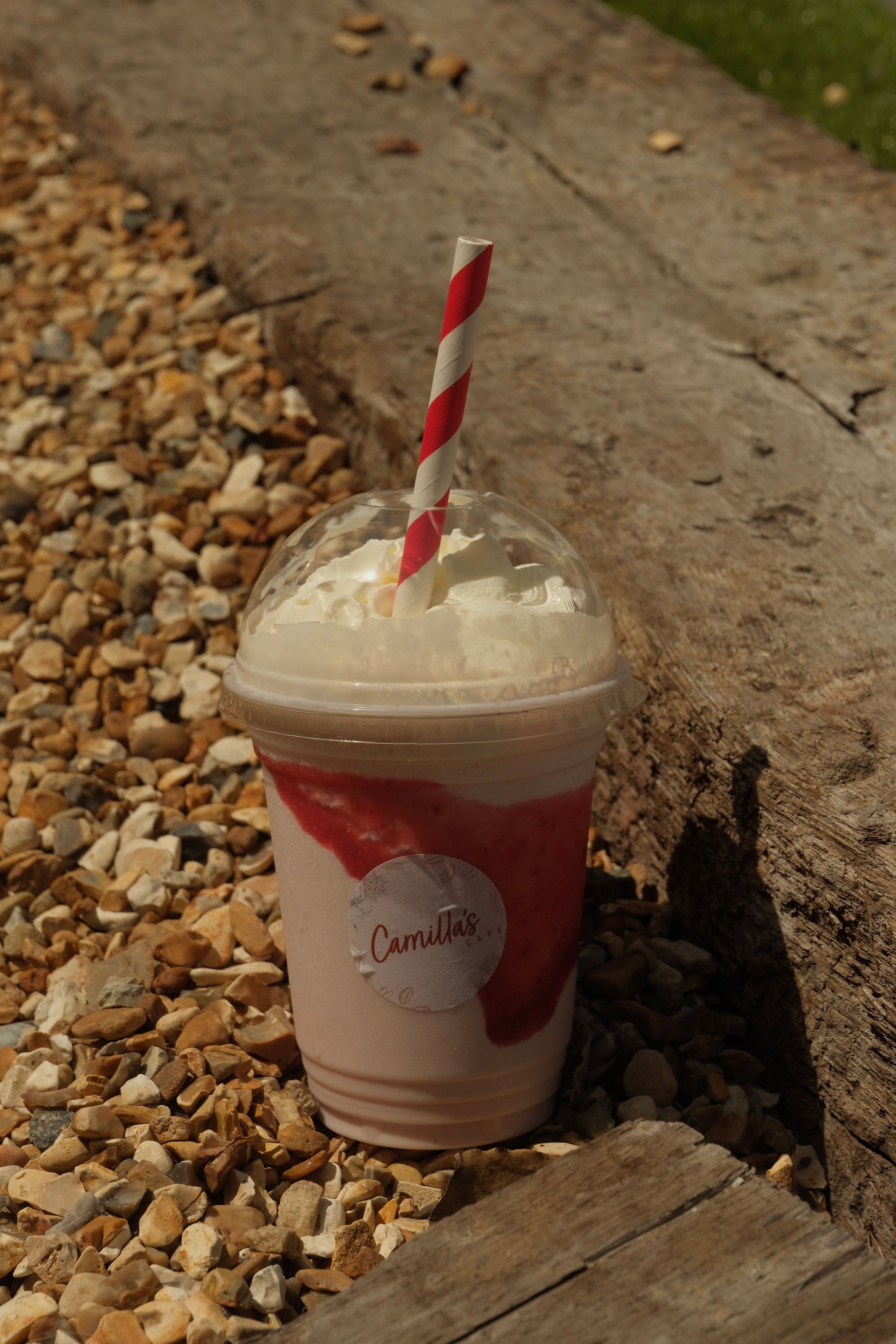 A strawberry milkshake with whipped cream, a red striped straw, and a "Camilla's" label sits on gravel next to a wooden beam.