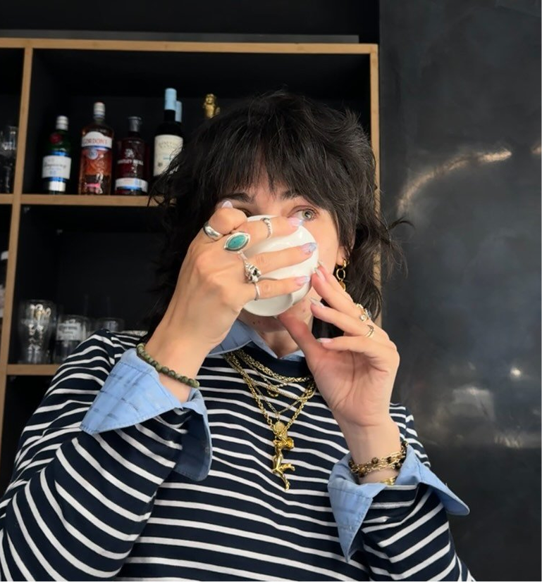 A person with dark shaggy hair, adorned with jewelry and a striped shirt, sips from a white cup.