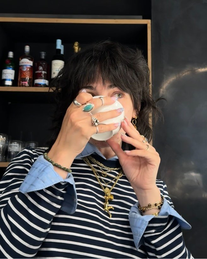 A person with dark shaggy hair, adorned with jewelry and a striped shirt, sips from a white cup.