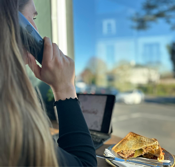 A person on a landline phone, seated by a window with a grilled sandwich and laptop on the table.