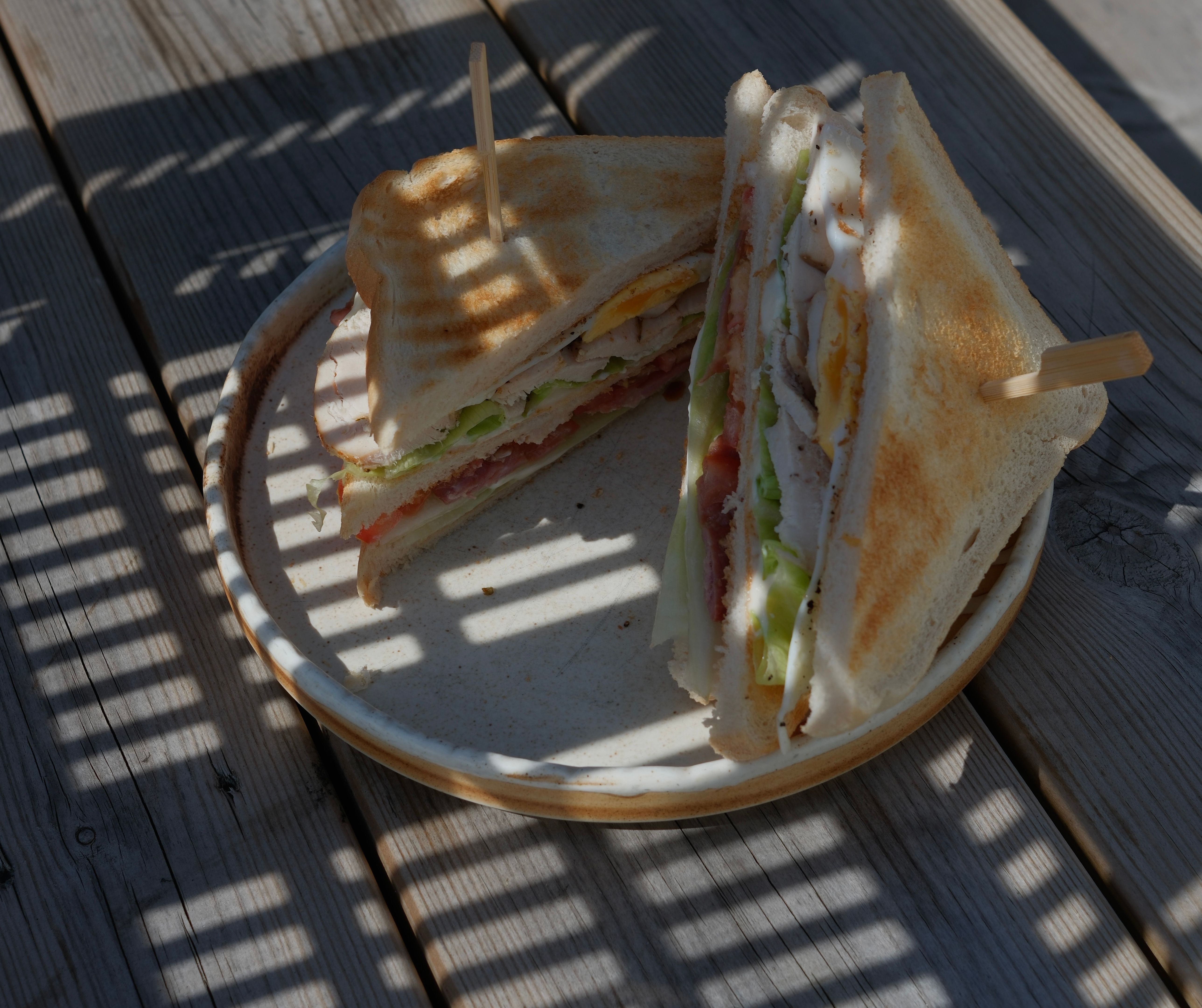 A club sandwich on a plate on a wooden table with striped shadows.