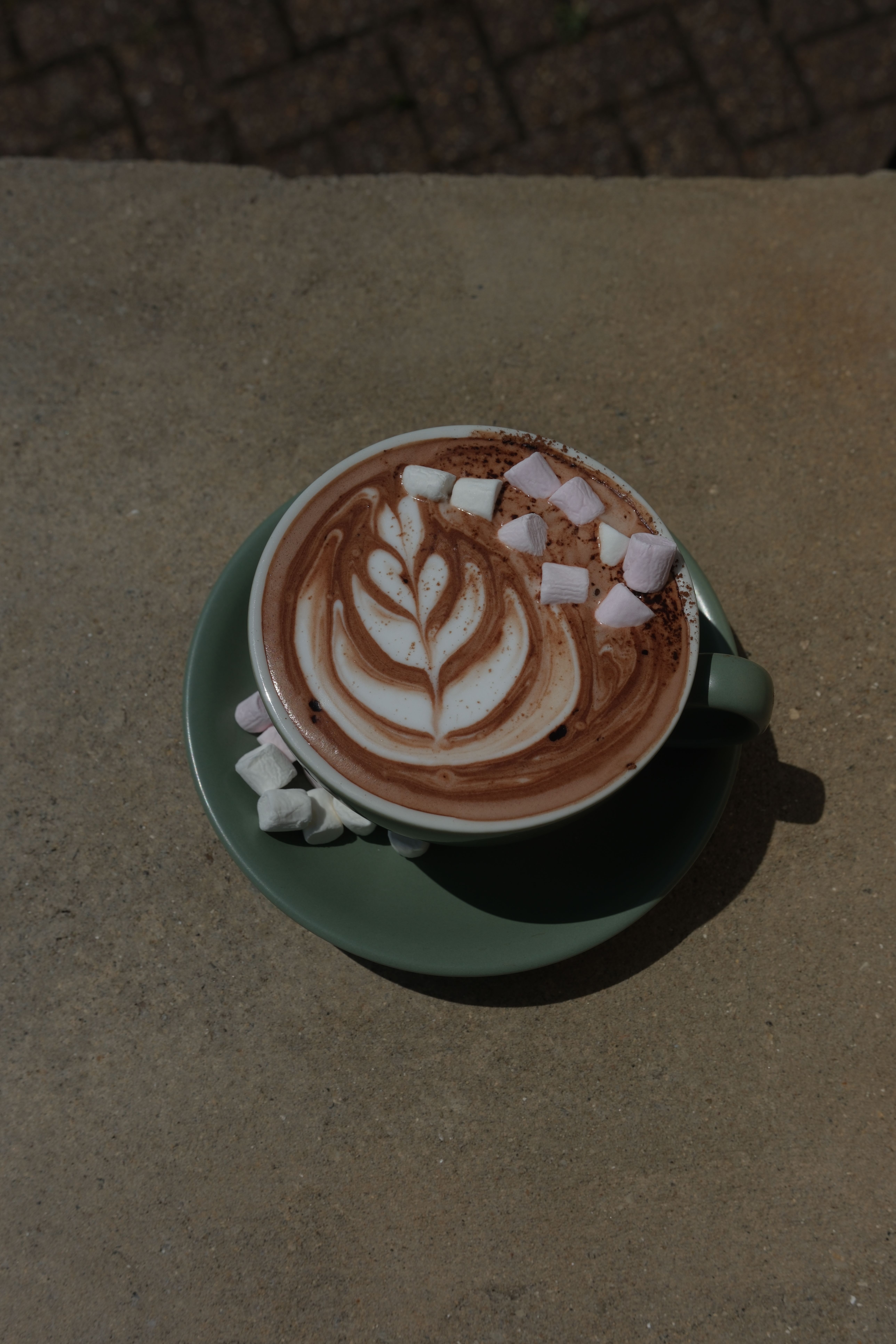 A hot chocolate with leaf latte art and pink and white marshmallows in a green cup and saucer on concrete.