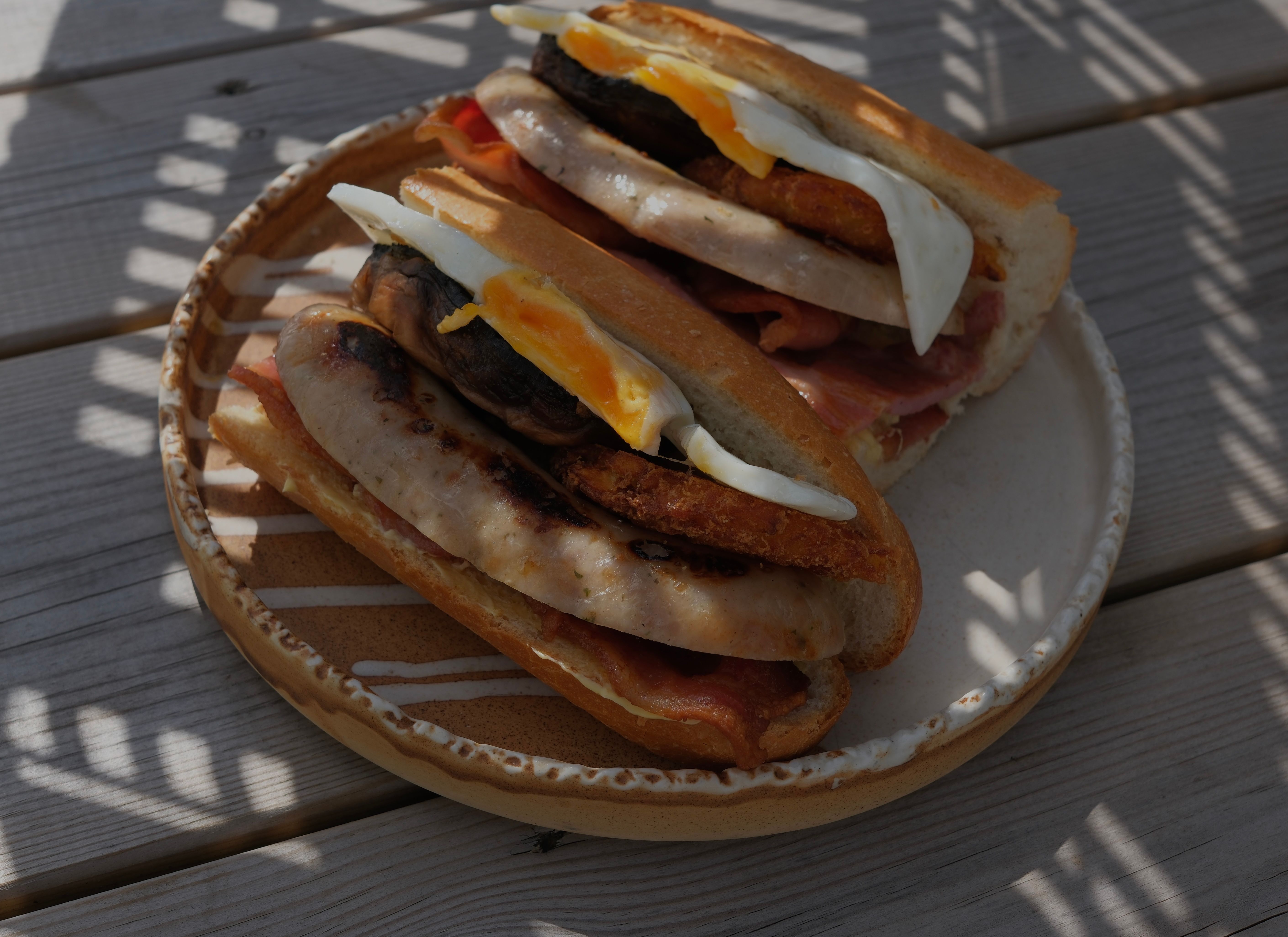 Two baguette sandwiches filled with sausage, bacon, and fried eggs, on a patterned plate set on a wooden table.