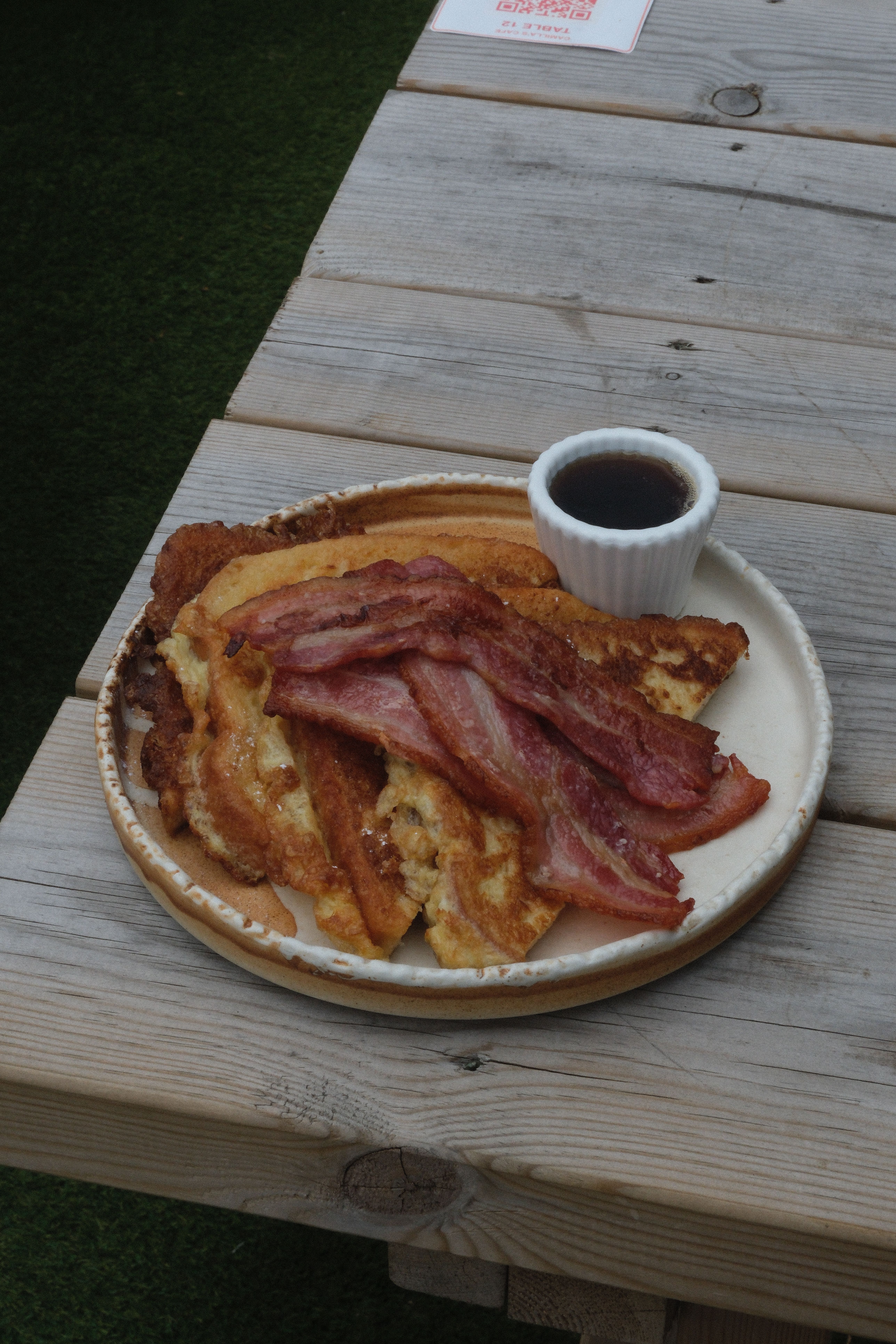 A plate of French toast topped with bacon, with a side cup of syrup, on a wooden table.
