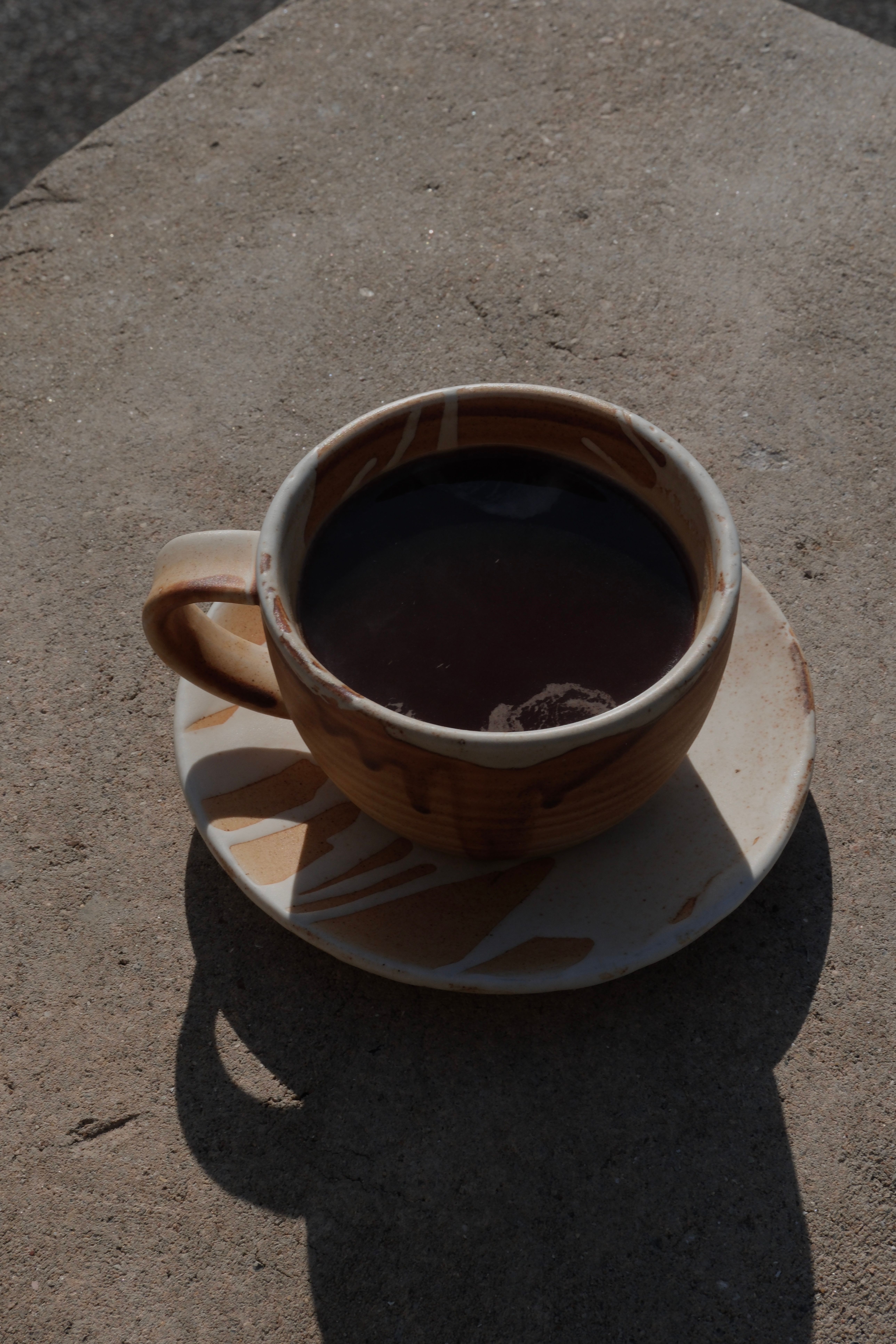 Rustic ceramic mug with dark liquid on a matching saucer, casting shadows on a concrete surface.