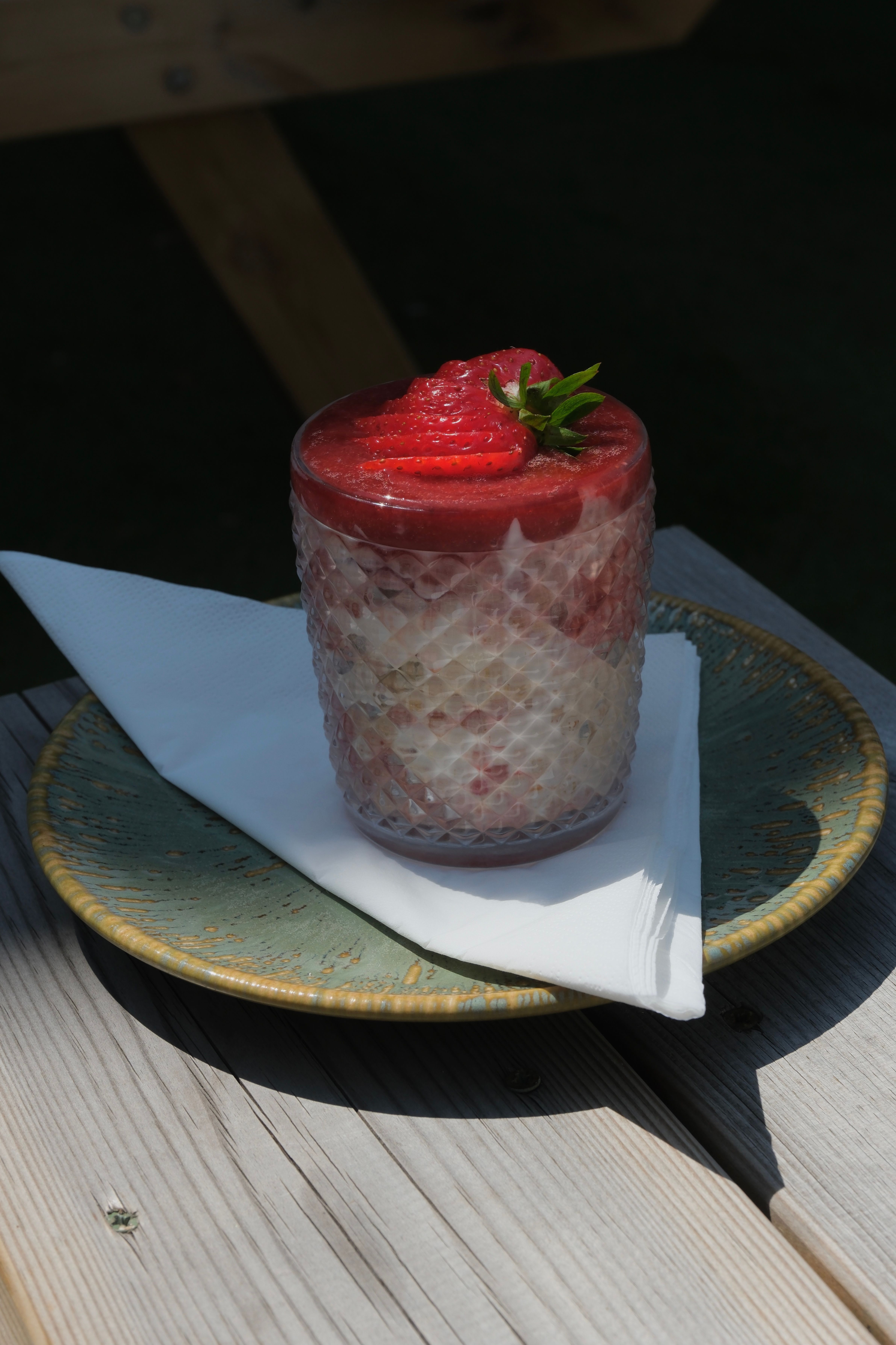 A layered strawberry dessert in a textured glass, topped with fresh strawberries and mint, on a green plate on a wooden table.