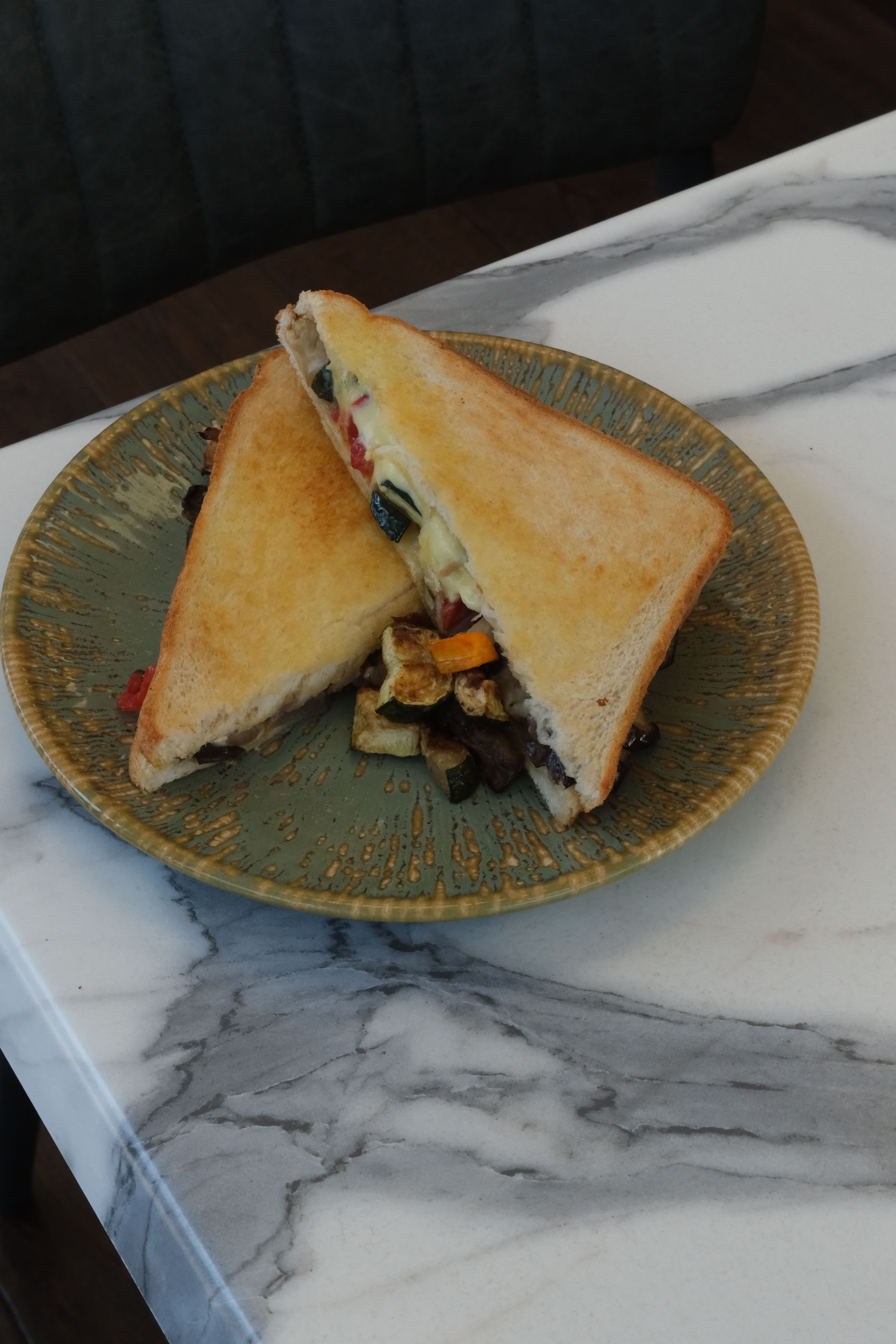 A toasted sandwich filled with melted cheese and grilled vegetables, cut diagonally, served on a green plate with a side of grilled vegetables.