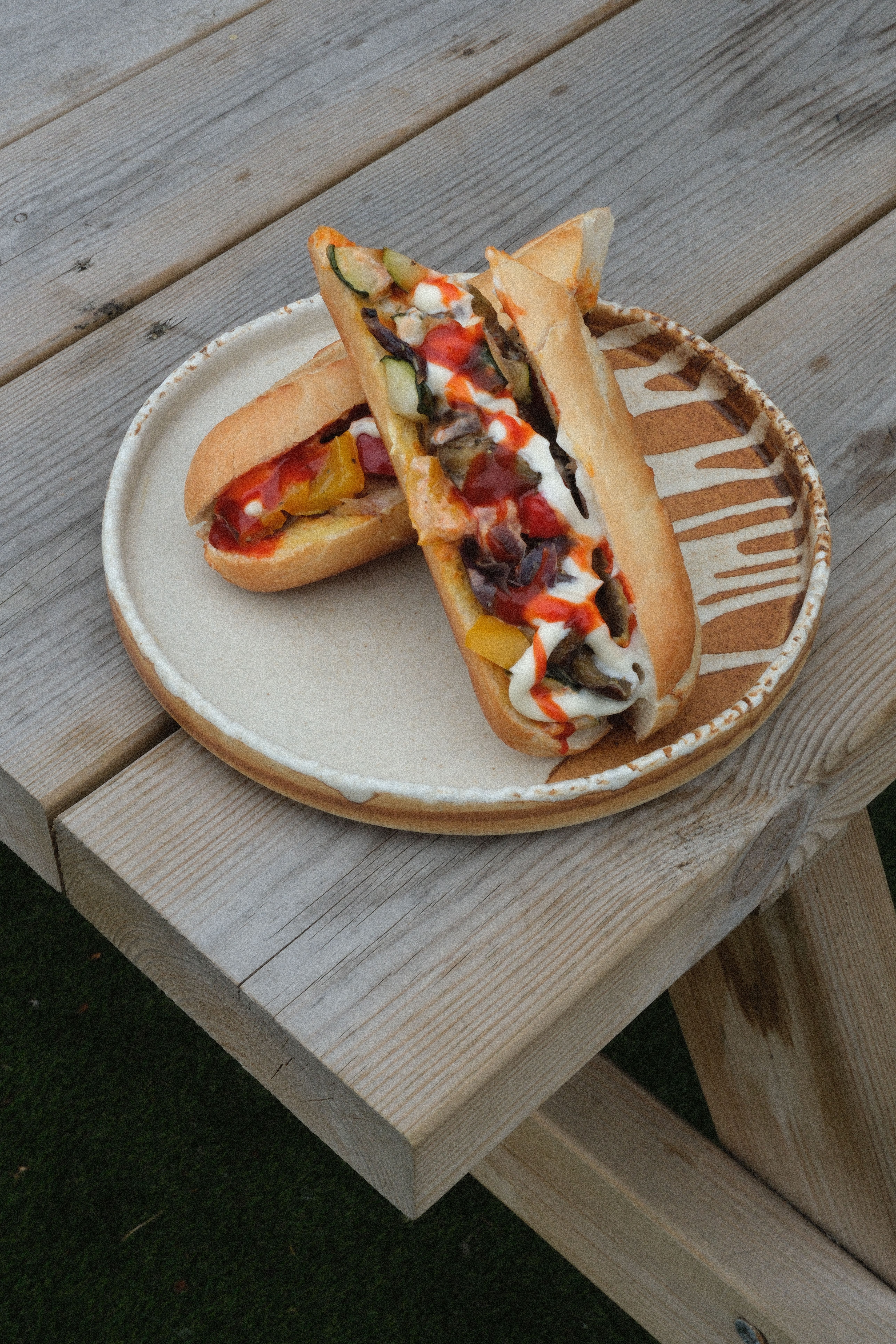 Two subs filled with grilled vegetables, white and red sauces, on a plate on a wooden picnic table.