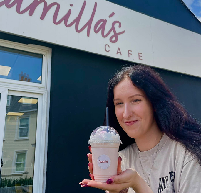 Smiling woman holds a pink milkshake with whipped cream in front of Camilla's Cafe.