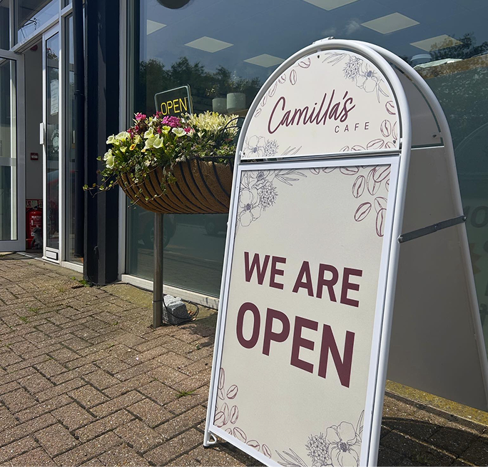 An A-frame sign for Camilla's Cafe reads "WE ARE OPEN" next to a hanging flower basket outside the cafe.