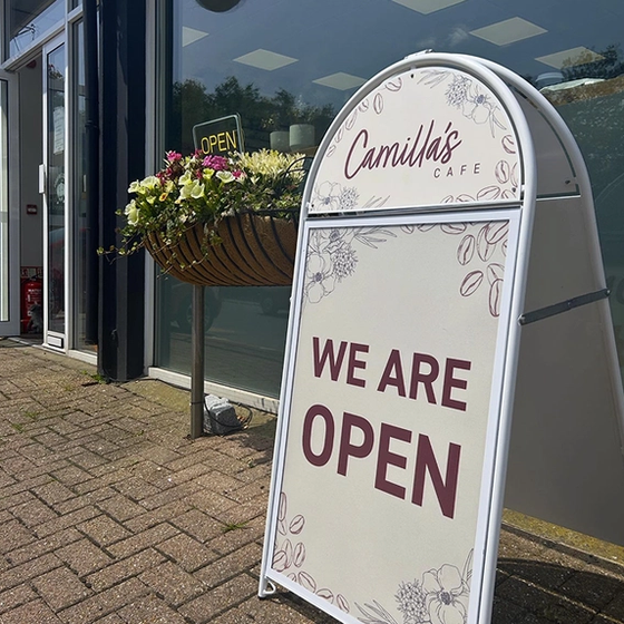 An A-frame sign for Camilla's Cafe reads "WE ARE OPEN" next to a hanging flower basket outside the cafe.