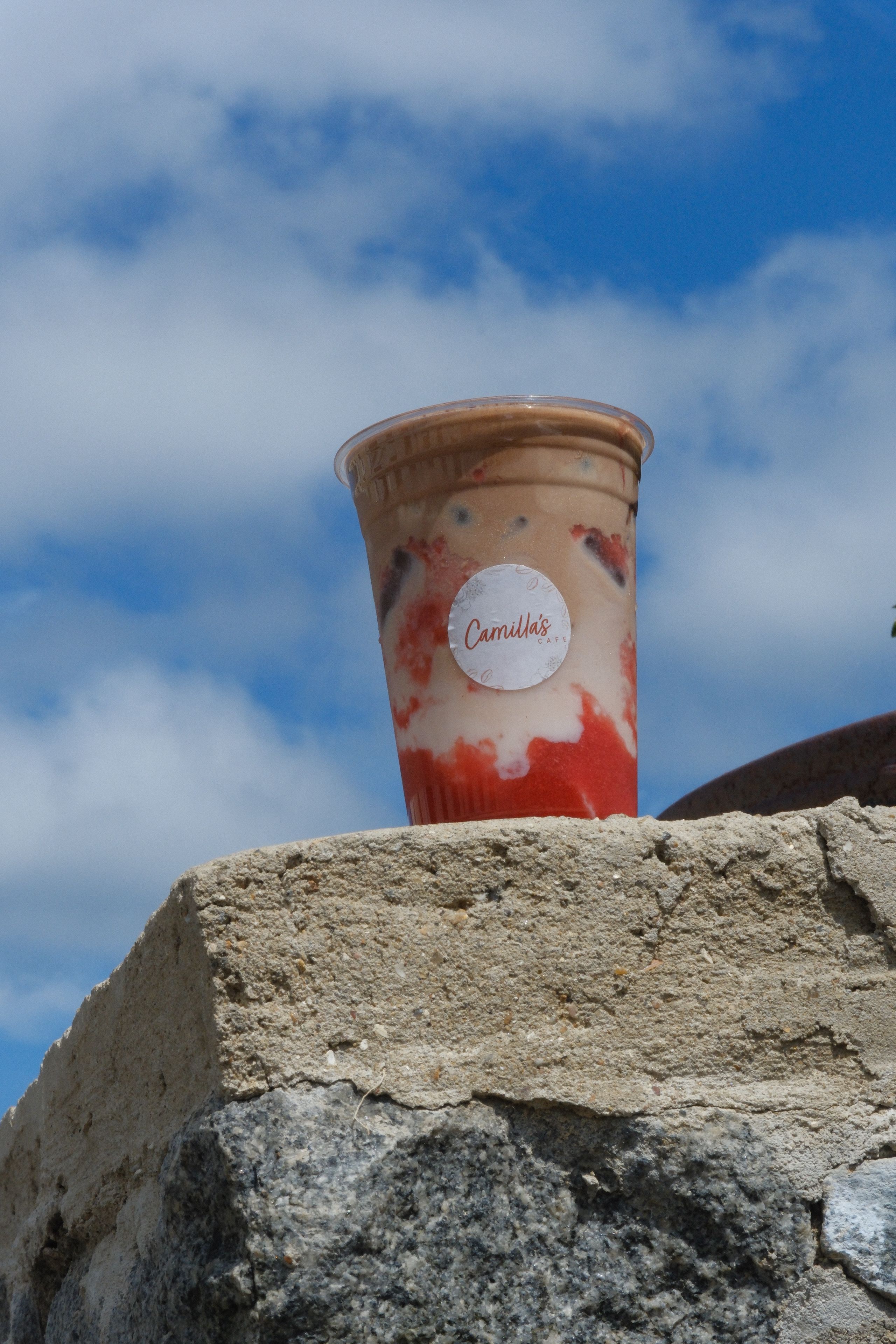 A Camilla's branded iced drink with strawberry puree and milk layers sits on a stone wall under a cloudy blue sky.