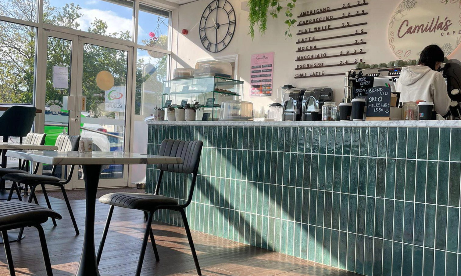 Interior of Camilla's Cafe with tables, a teal tiled counter, menu, and a barista, illuminated by sunlight from large windows.