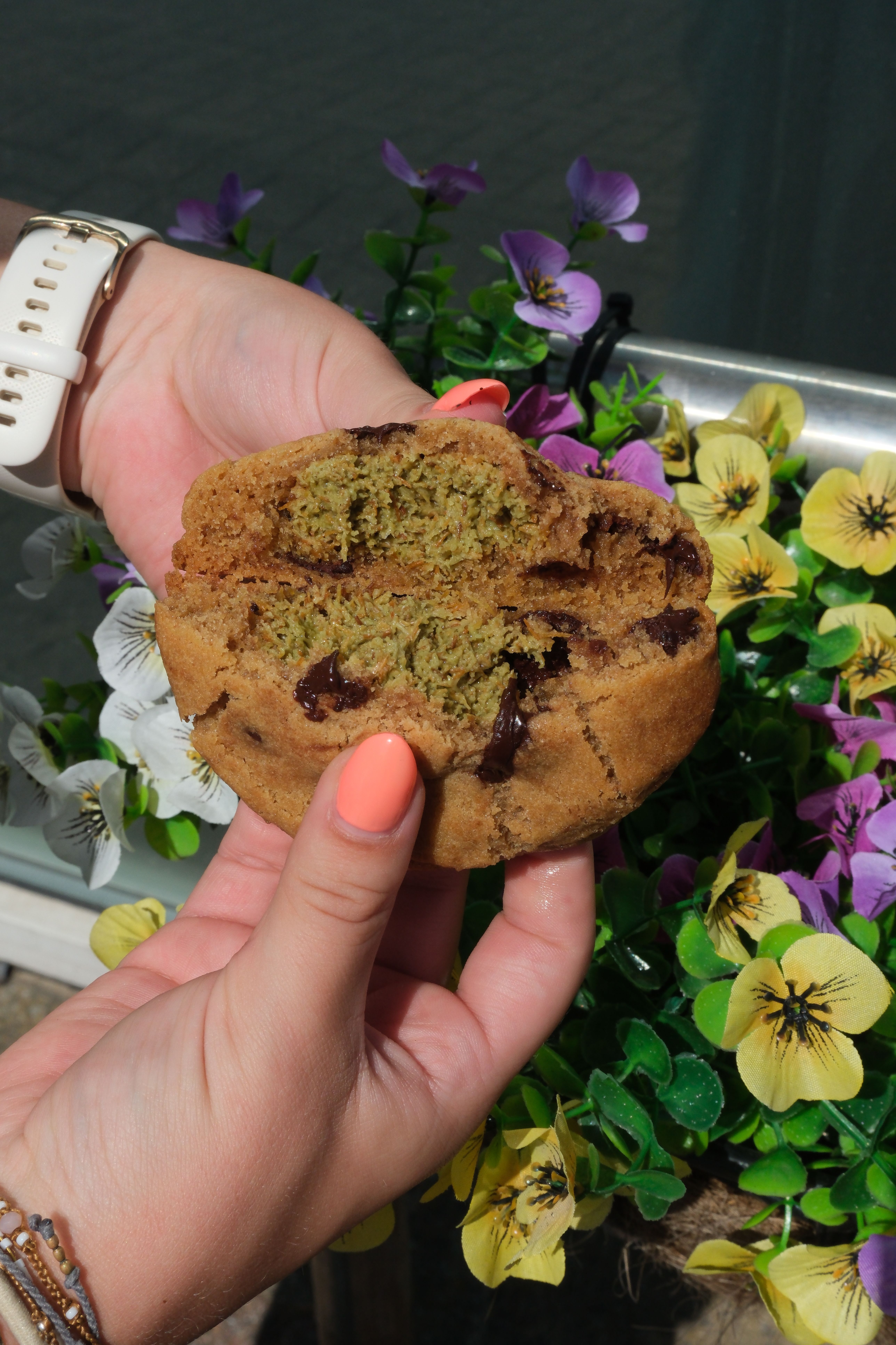 A person's hands hold a chocolate chip cookie broken in half, revealing a green filling, with colorful flowers in the background.