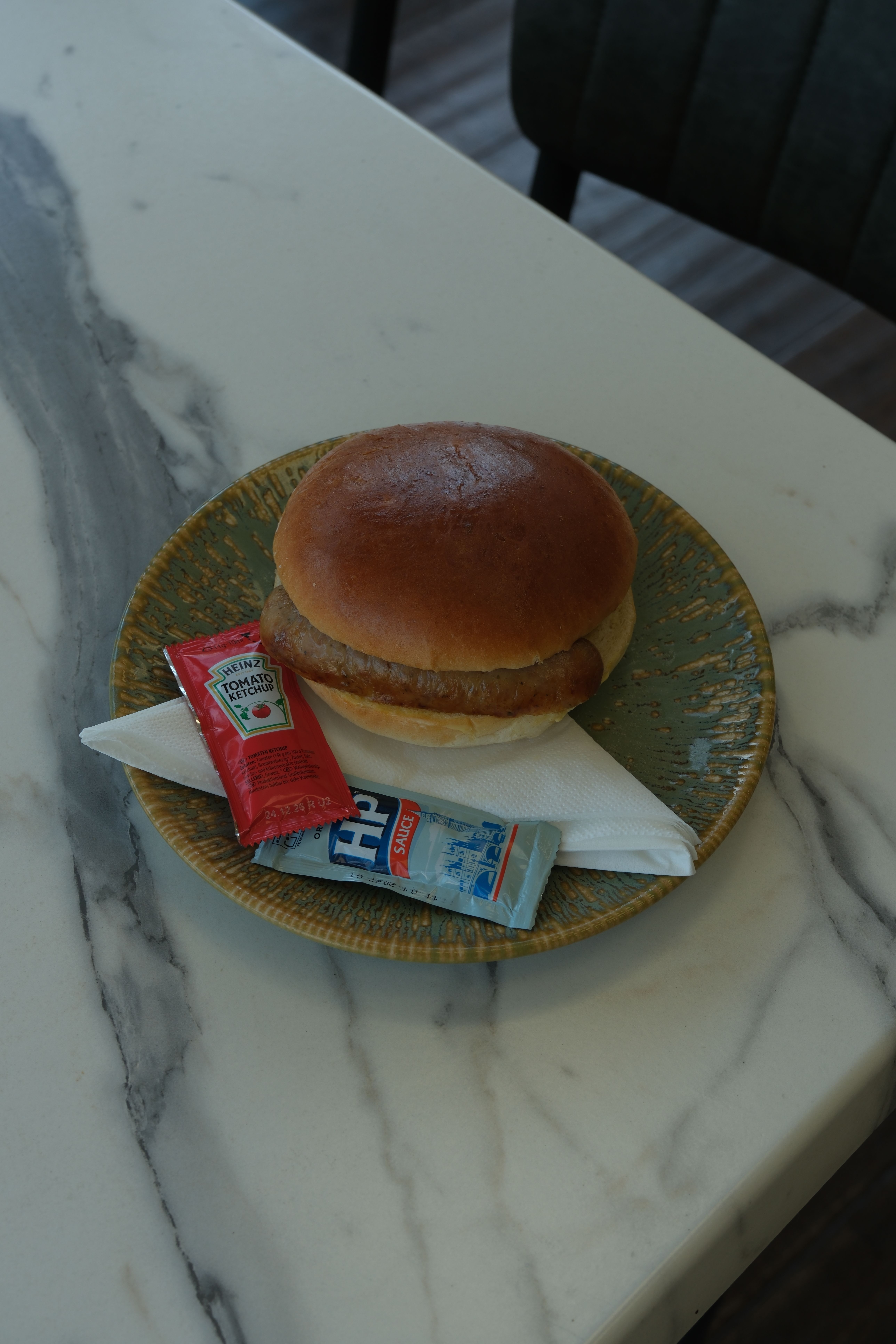 A sausage bun on a green plate with Heinz Ketchup and HP Sauce packets on a marble table.