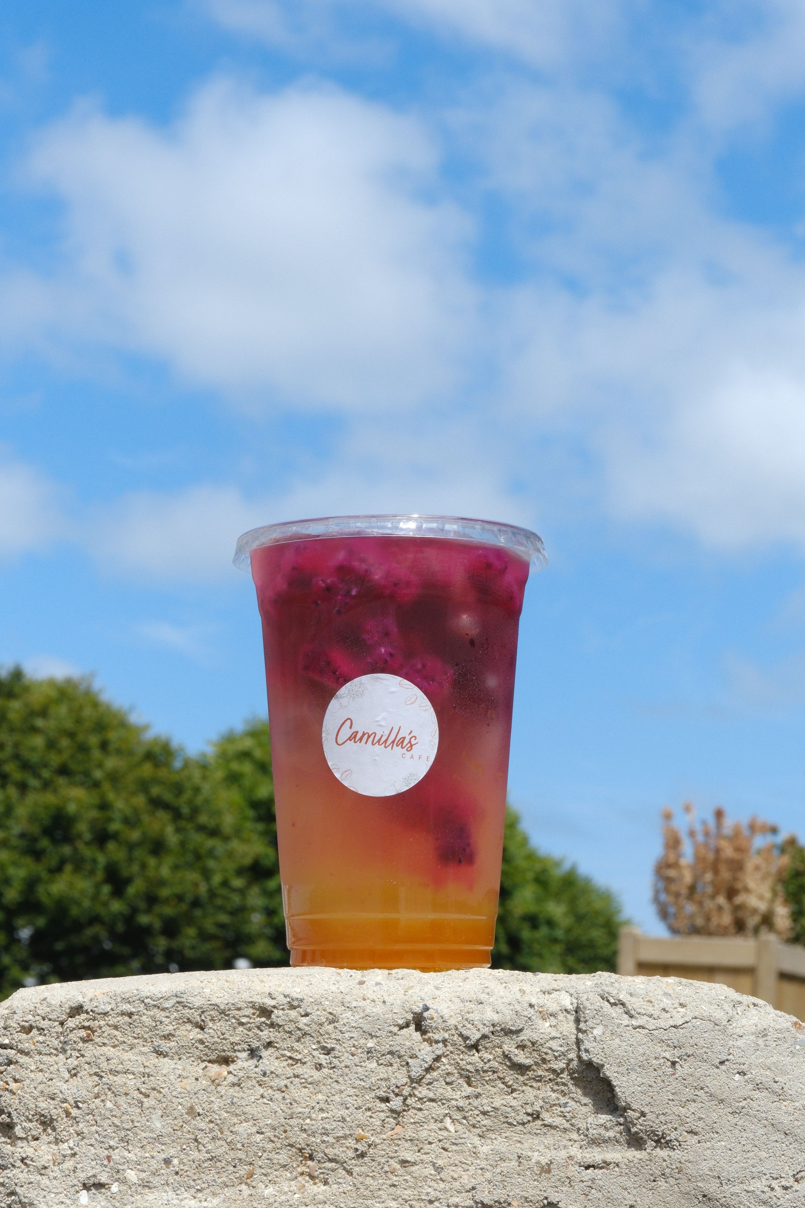 A clear cup with a yellow and pink layered drink, containing dragon fruit chunks and a 'Camilla's' label, sits on a stone wall under a blue sky.