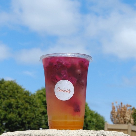 A clear cup with a yellow and pink layered drink, containing dragon fruit chunks and a 'Camilla's' label, sits on a stone wall under a blue sky.