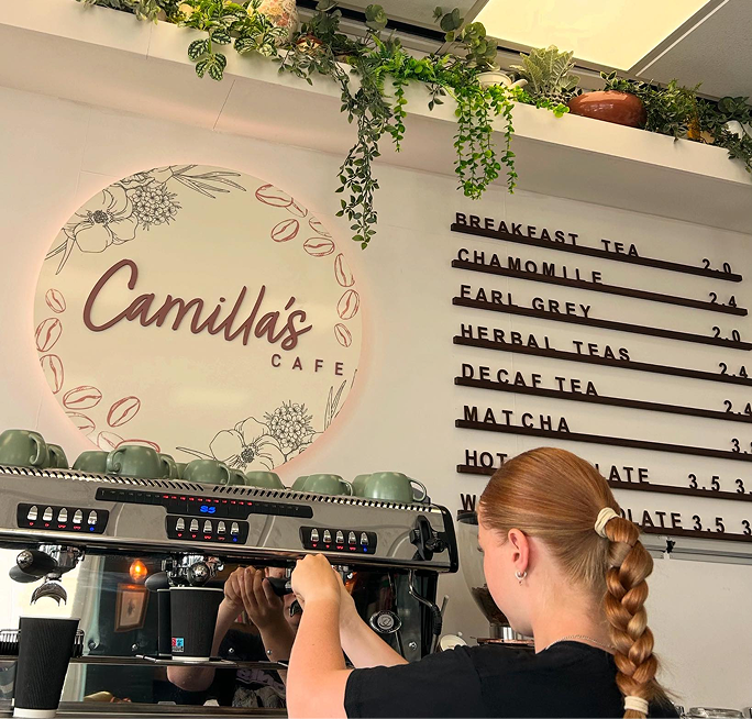 A barista operates an espresso machine at Camilla's Cafe, which features a circular sign and a menu board listing teas.