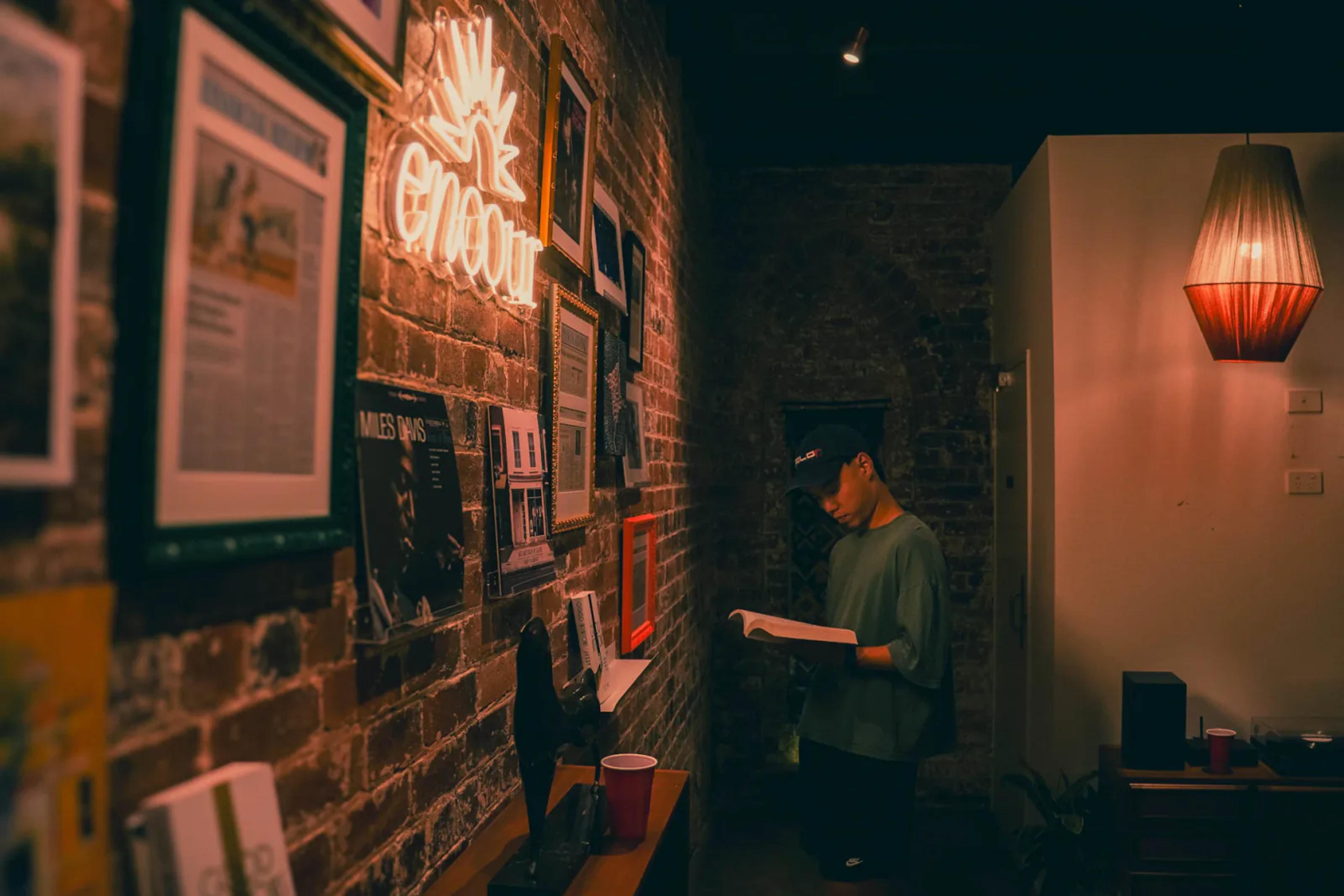 A person reading by a brick wall of framed clippings and artwork at Broad Church, lit by a glowing "encour" neon sign