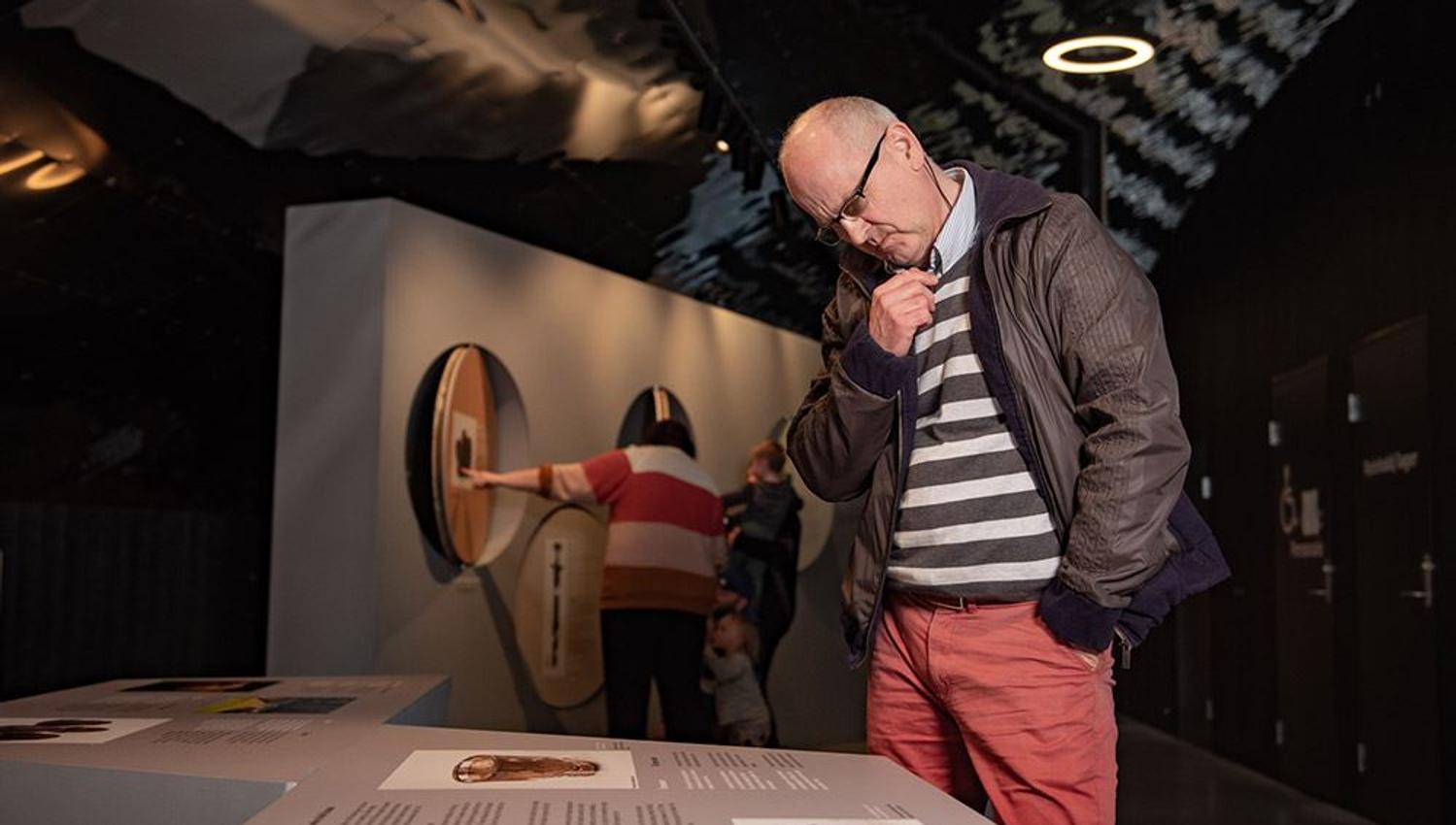 Besøkande les informasjon og lærer om vikingane på Sagastad. Informasjonstavle i ei mørk museumutstilling, med andre besøkande i bakgrunnen..