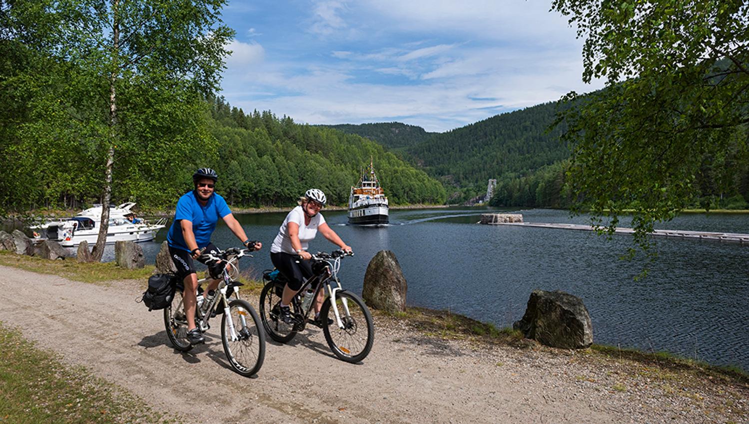 couple is bicycling along the Telemark Canal with MS Victoria in the background