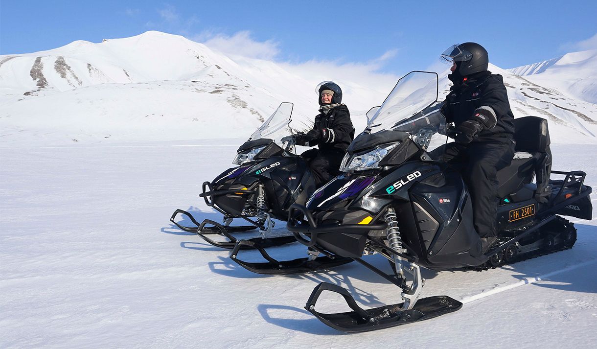 To personer i snøscooterutstyr på elektriske snøscootere som smiler til hverandre, med et snødekt landskap rundt seg