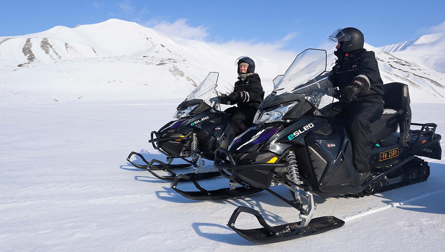 To personer i snøscooterutstyr på elektriske snøscootere som smiler til hverandre, med et snødekt landskap rundt seg
