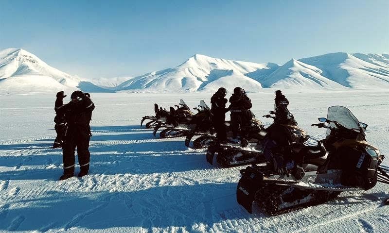 Sunny day with mountains in the background. In front it's a line of snowmobiles with people standing around.