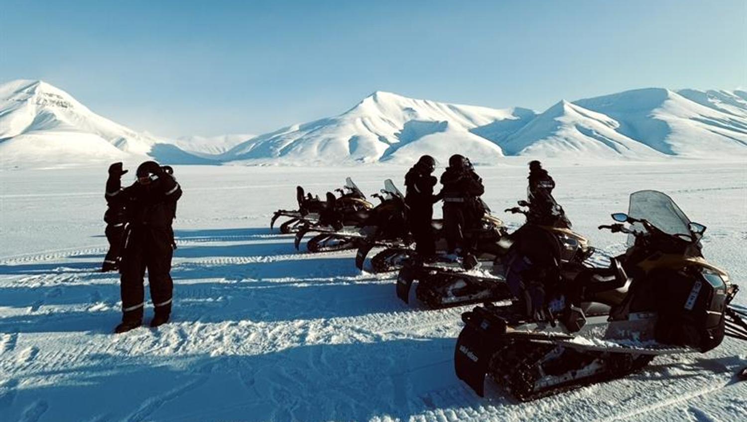 Sunny day with mountains in the background. In front it's a line of snowmobiles with people standing around.